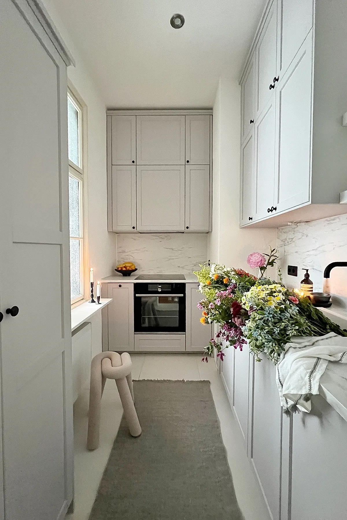narrow white kitchen with tall cabinets, marble backsplash, built-in oven, black faucet, window on left side, bouquet of mixed flowers on counter