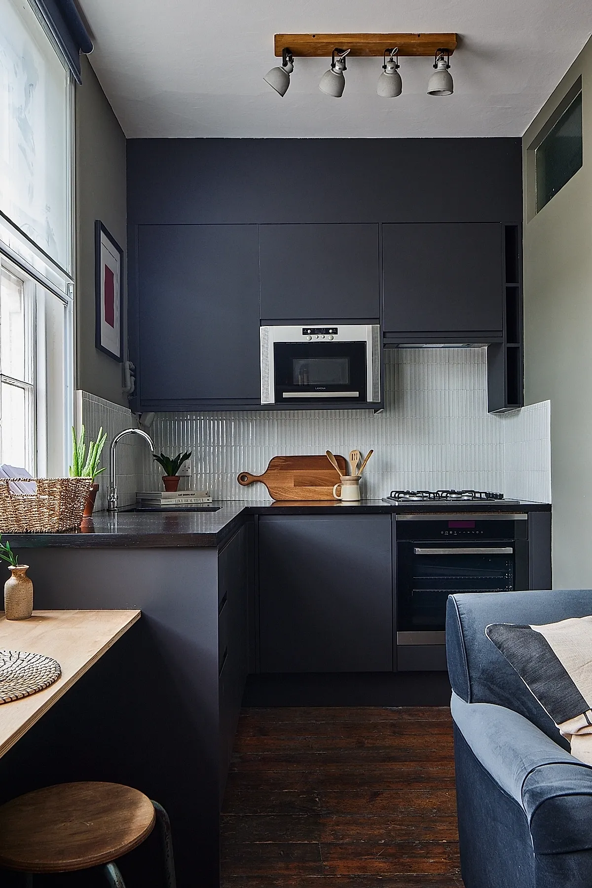 small L-shaped kitchen with dark gray cabinets, black countertop, stainless steel microwave and oven, wooden cutting board and utensils, white vertical tile backsplash, wood ceiling light fixture