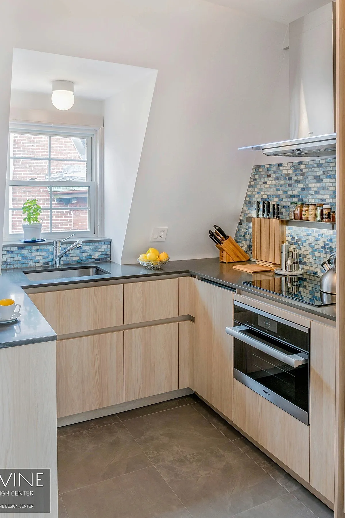 small kitchen with light wood cabinets, gray countertops, blue mosaic tile backsplash with built-in spice shelf, stainless steel oven and range hood, window above sink