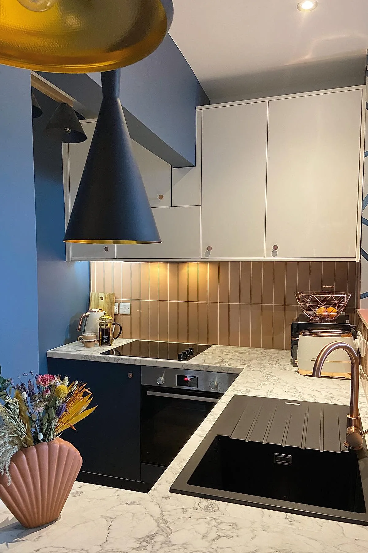 small kitchen with blue walls, white upper cabinets, black lower cabinets, marble countertops, brown vertical tile backsplash, black sink and faucet, under cabinet lighting, brass pendant lamp