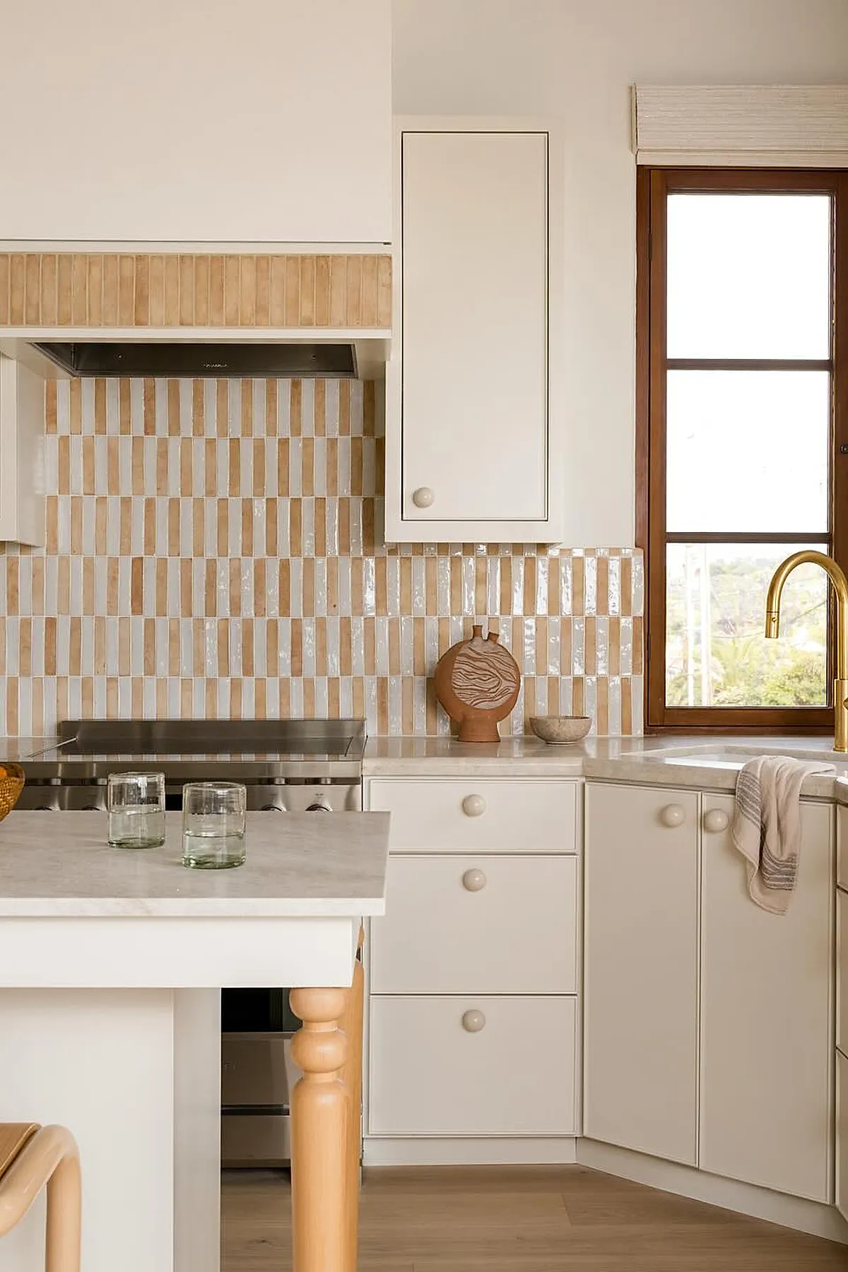 small kitchen with cream cabinets, vertical beige and white striped tile backsplash, brass faucet, wooden window frame, light countertop, minimal decor