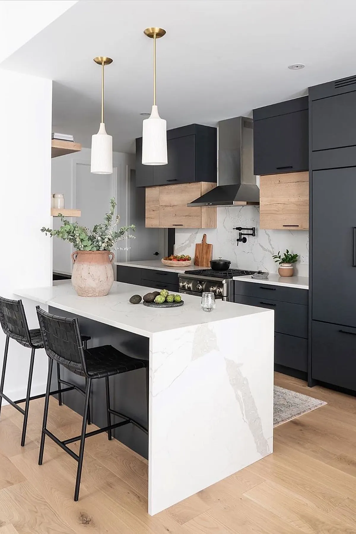 small kitchen with dark gray lower cabinets, light wood upper cabinets, marble waterfall island with black base, brass pendant lights, stainless steel range hood, light wood flooring