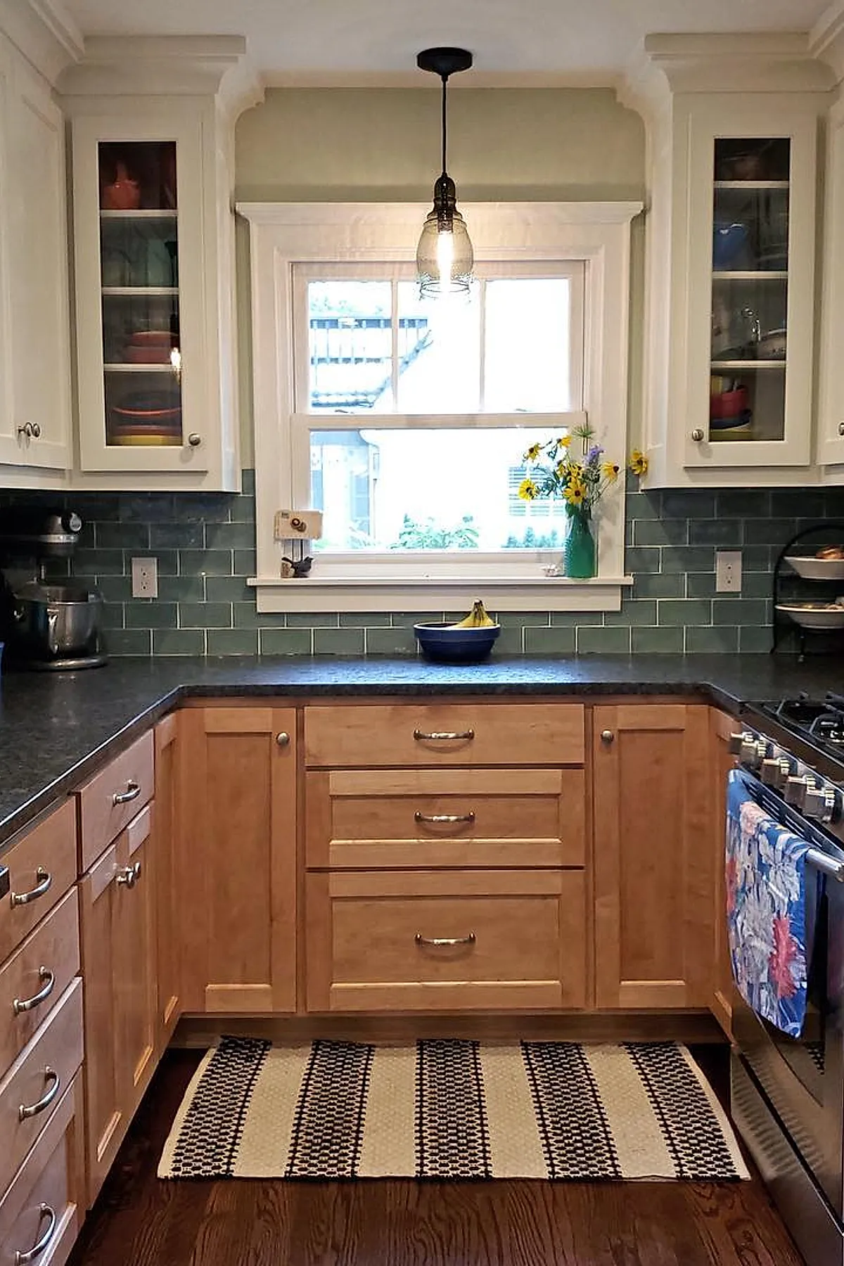 small kitchen with white upper cabinets, wood lower cabinets, green subway tile backsplash, black countertop, striped black and beige woven rug on dark wood floor, window above counter with flowers in vase