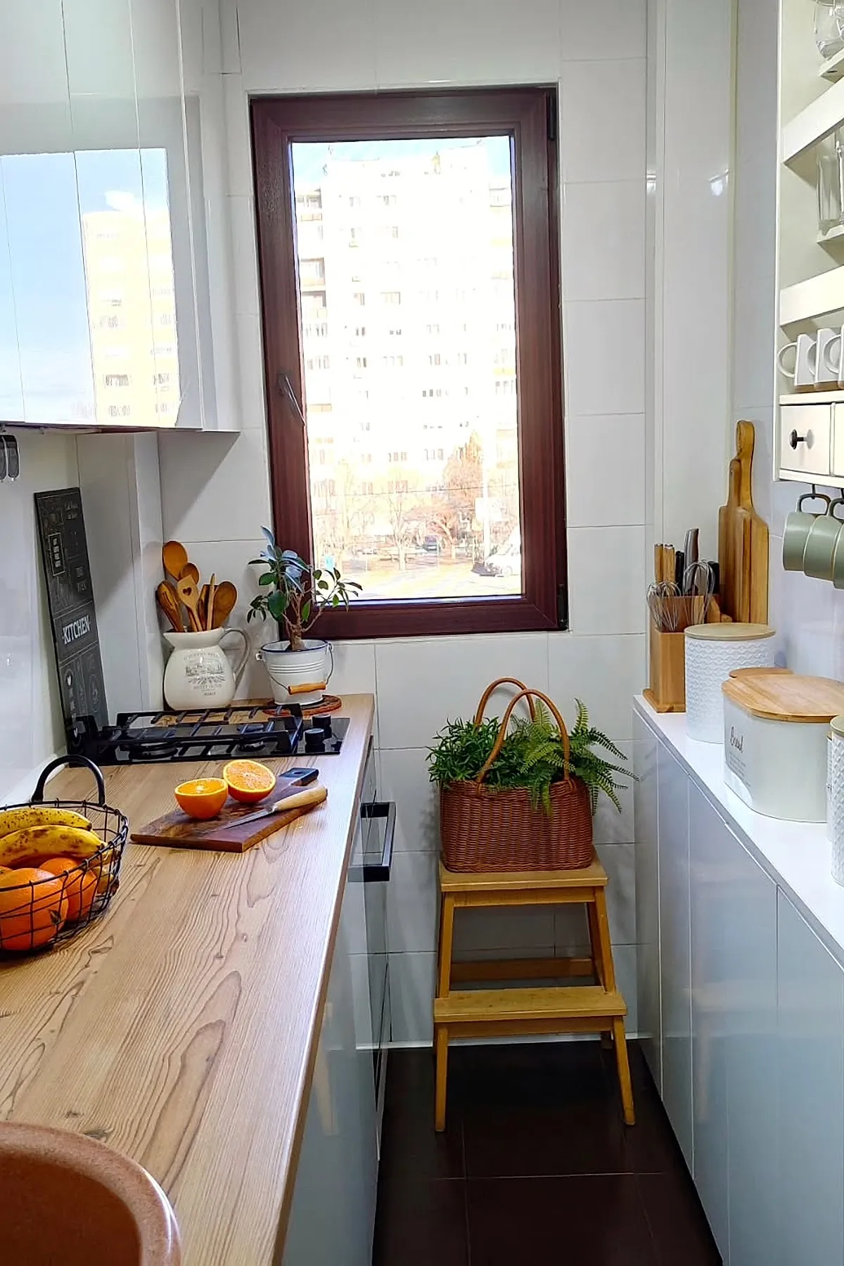 small narrow kitchen with light wood countertops, white cabinets, dark floor tiles, wooden window frame, potted plants on stool and countertop, fruit basket on counter
