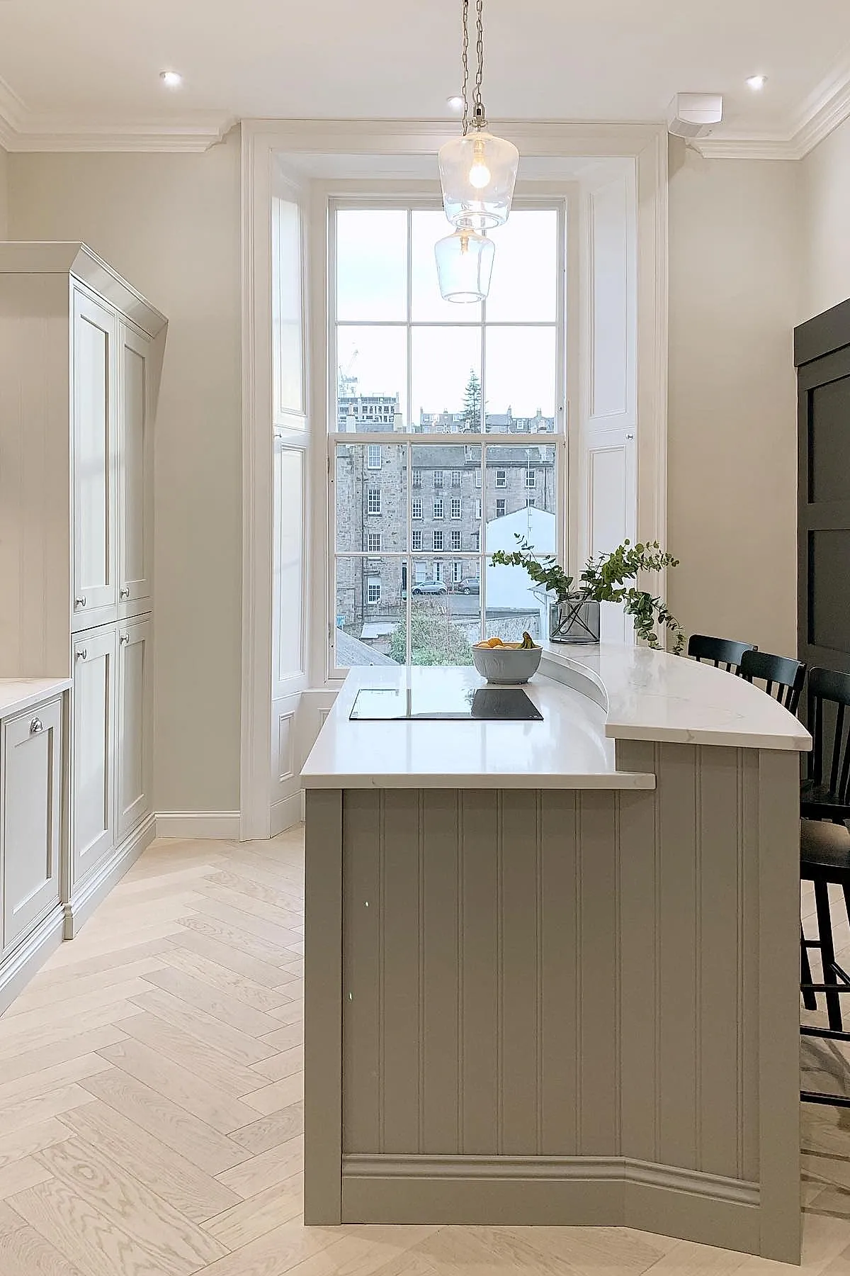 small kitchen with curved beige island featuring white countertop, large tall window behind, light wood herringbone floor, pendant lights, black chairs, and greenery on counter