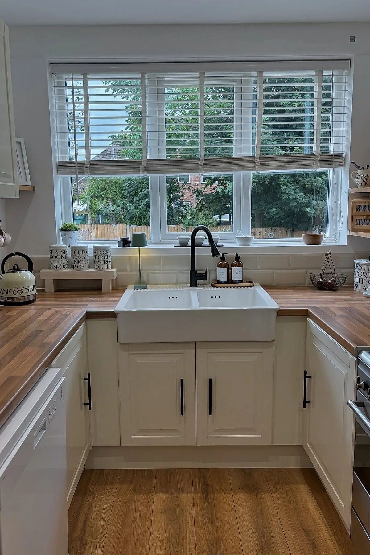 small U-shaped kitchen with white cabinets, wood countertops, double basin white sink, black faucet, large window with white blinds and green outdoor view