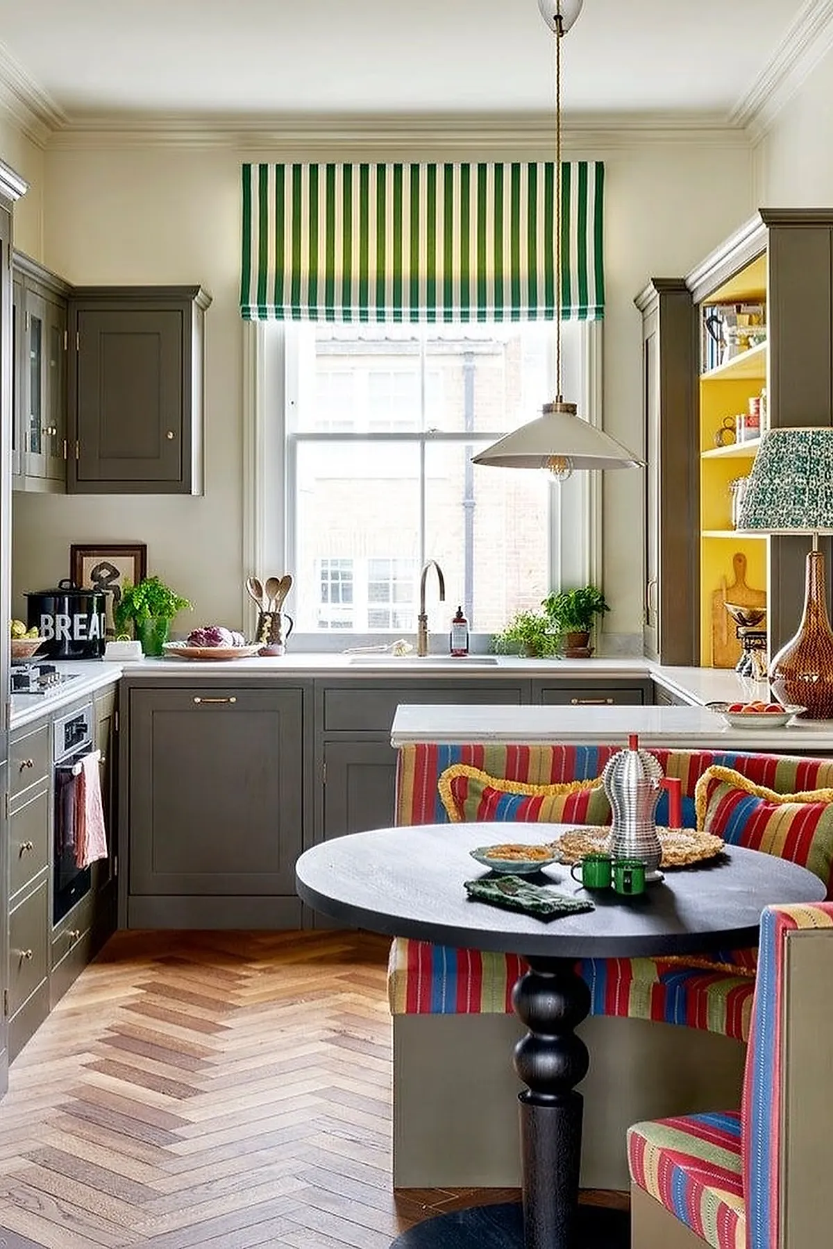 small kitchen with gray cabinets, white countertops, colorful striped banquette seating around black round table, green and white striped window blind above sink, wood herringbone floor