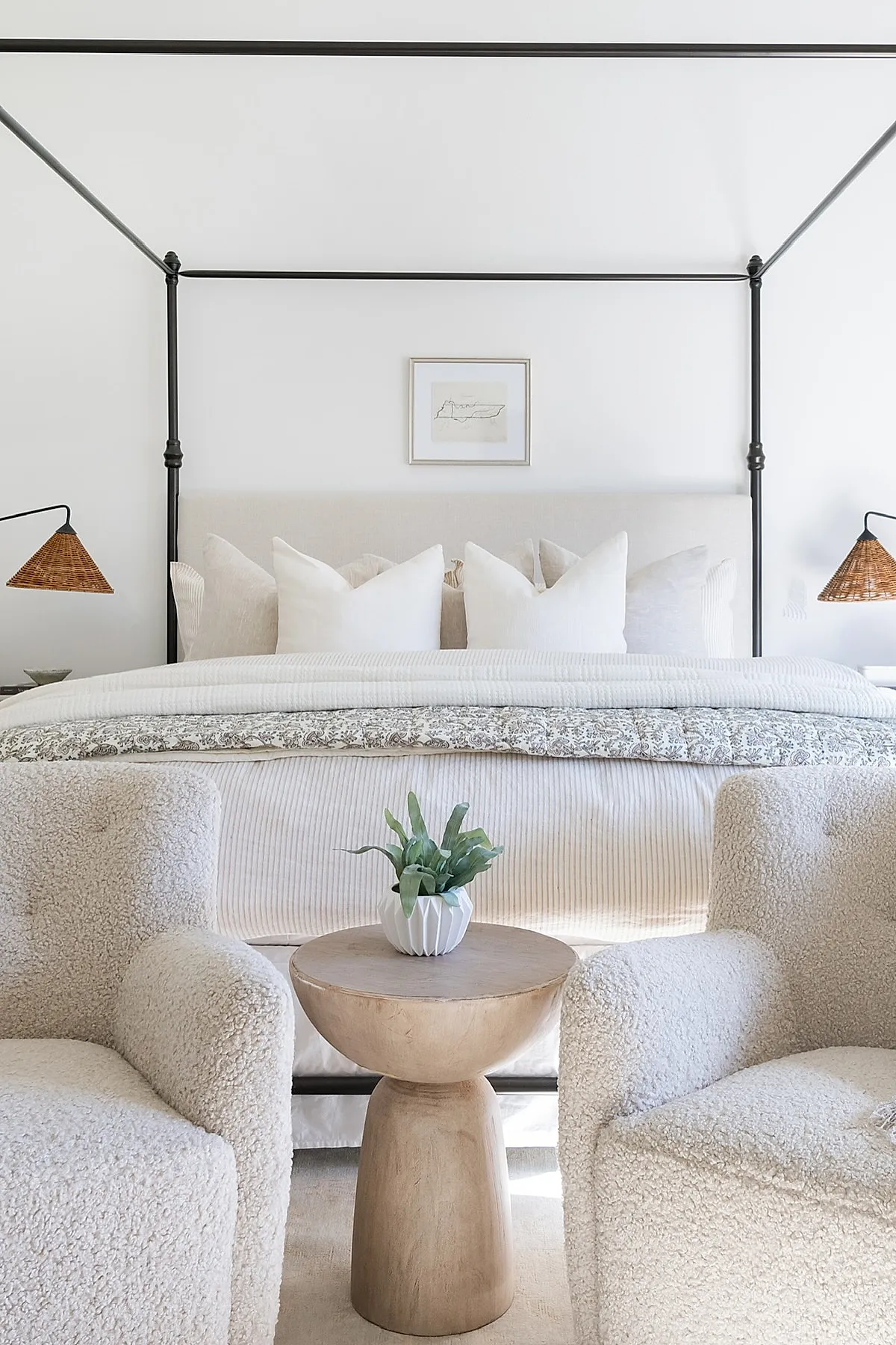 Bedroom featuring a black metal canopy bed with cream bedding, two off-white textured armchairs, a small light wood side table with a potted green plant, and white walls.