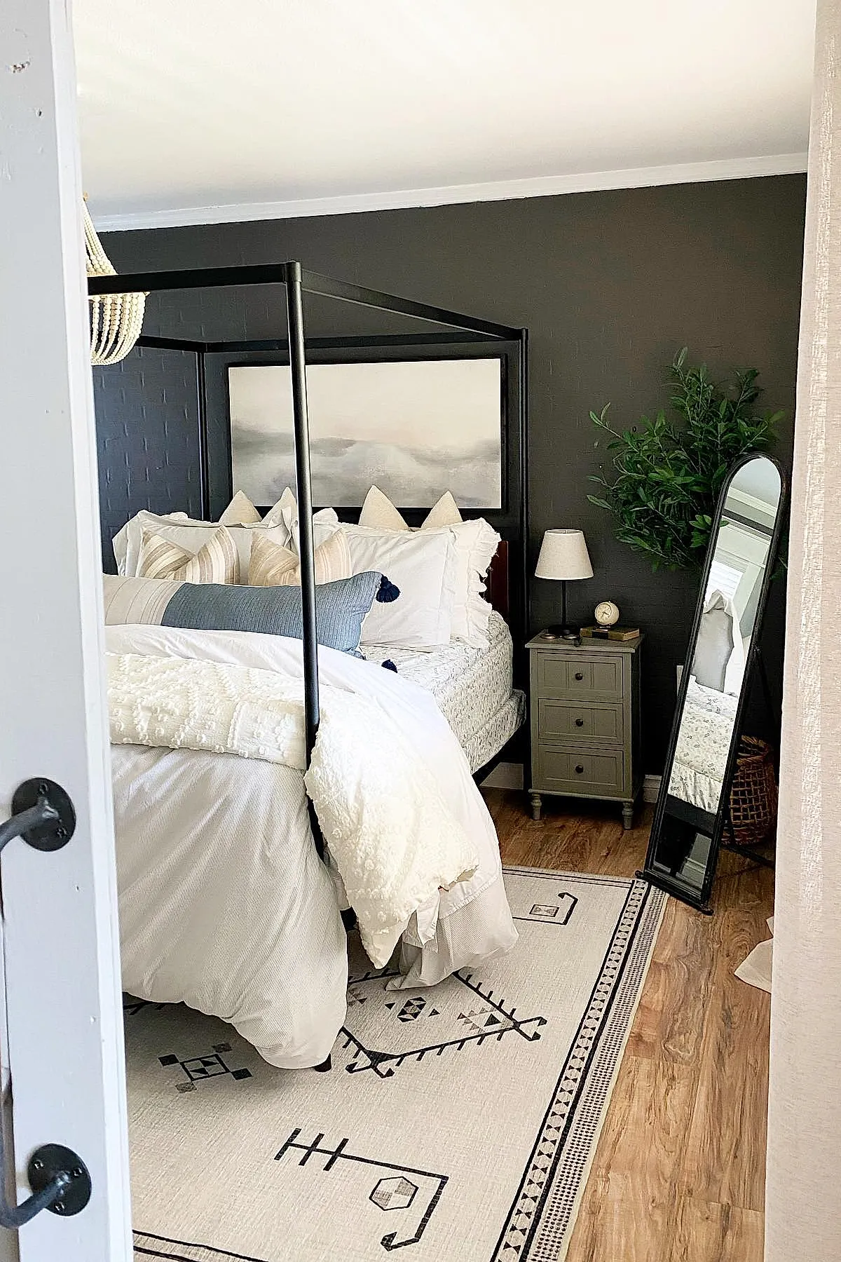 Bedroom featuring a black metal canopy bed with white and beige bedding, dark charcoal painted brick accent wall, light patterned area rug on wood floor, gray nightstand with lamp, tall green plant next to a black framed full-length mirror.