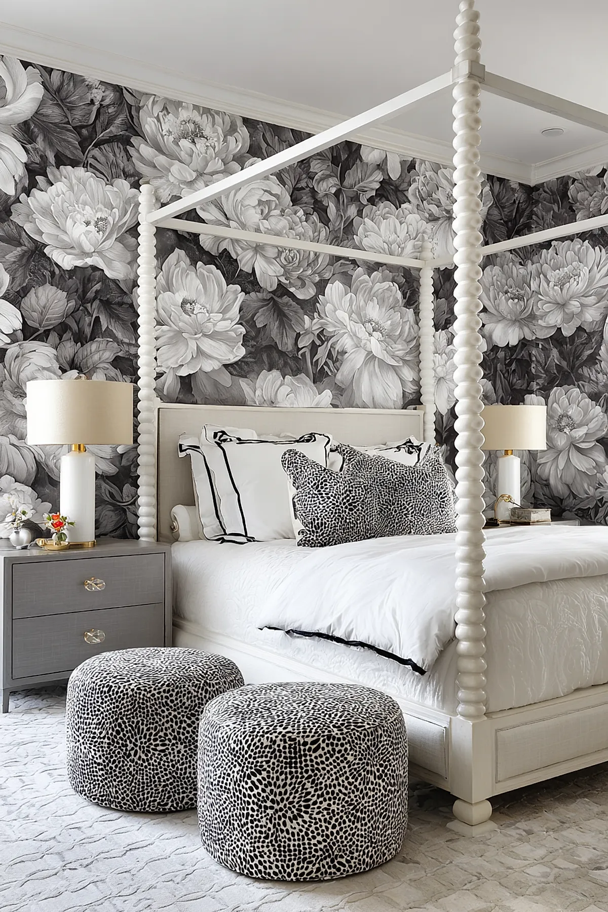 White canopy bed with turned posts in front of black and white large floral wallpaper, featuring white bedding with patterned pillows, two spotted round ottomans at foot, gray nightstands each holding a lamp.