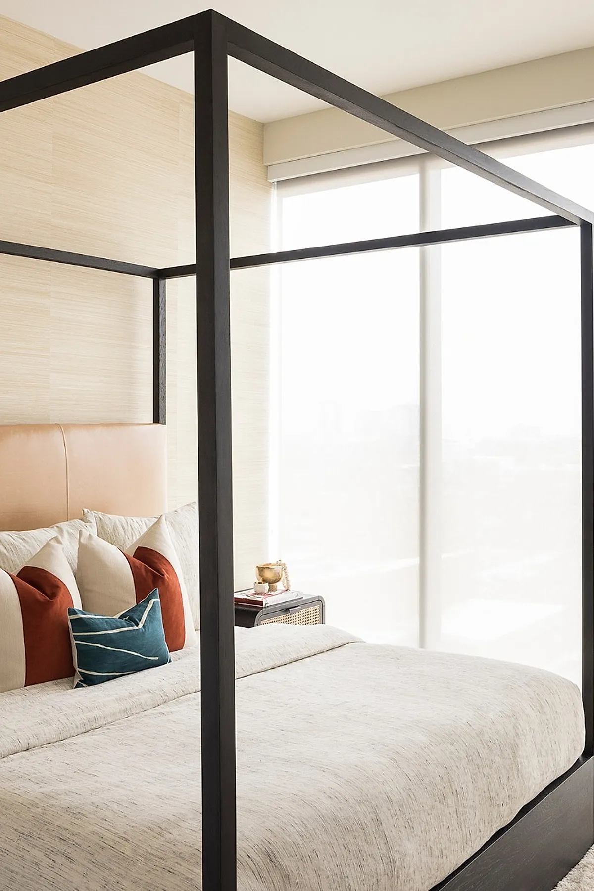 Bedroom featuring a modern black metal canopy bed with beige bedding, tan leather headboard, white pillows accented by terracotta and blue velvet decorative pillows, next to large sheer window panels.