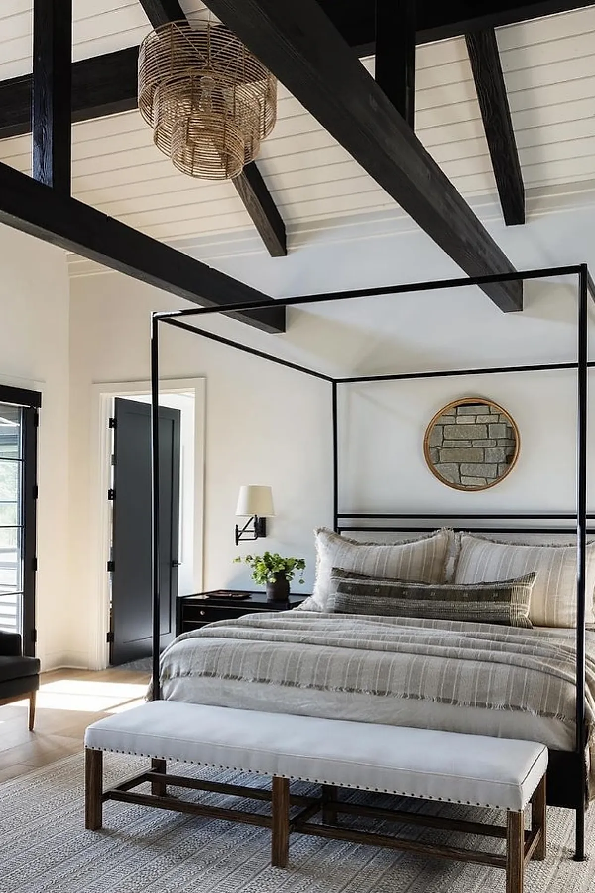 Bedroom with black metal canopy bed, neutral striped linen bedding, gray textured rug, wooden bench with white cushion at foot of bed, round mirror with wood frame on white wall, exposed dark wood ceiling beams.