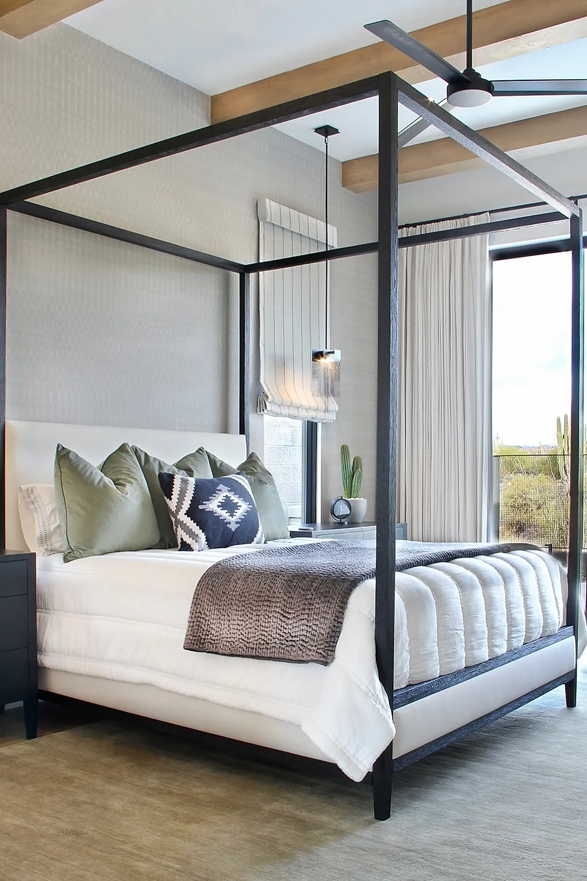 Bedroom featuring black metal canopy bed with white bedding, olive green and navy geometric pillows, gray textured throw blanket, small side table with potted cactus, light walls with striped Roman shade, wood ceiling beams.