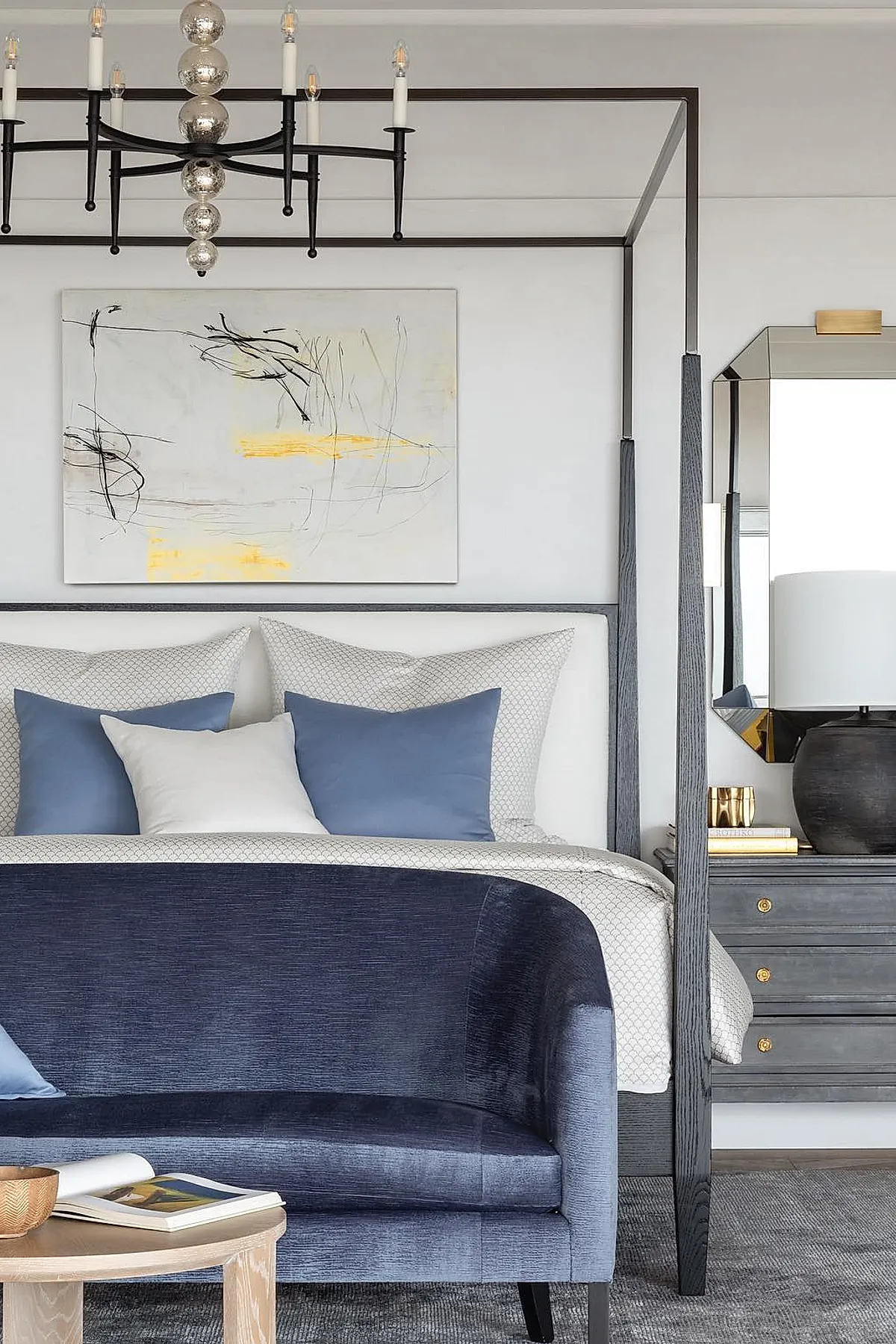 Bedroom featuring gray wood canopy bed with white and patterned bedding accented by blue pillows, deep blue velvet curved sofa at foot of bed, abstract wall art above headboard, dark wooden nightstand with black lamp.