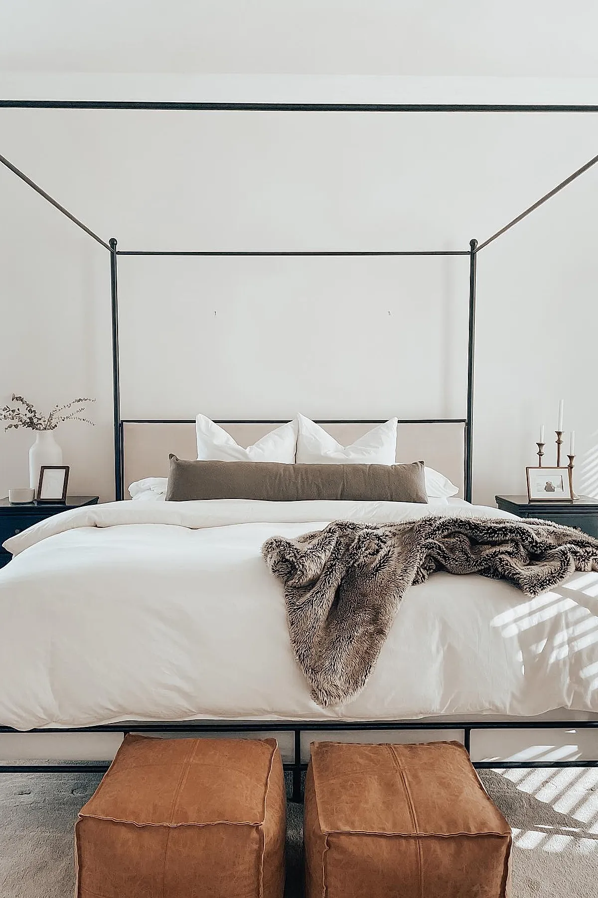 Bedroom featuring black metal canopy bed with white bedding, taupe lumbar pillow, faux fur throw blanket draped over the bed, two brown leather poufs at foot of bed, black nightstands on each side with minimal decor