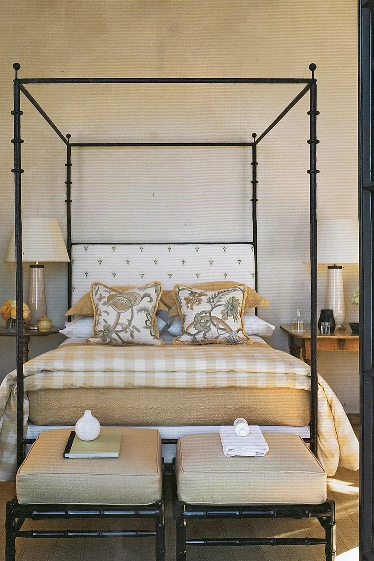 Black metal canopy bed with white headboard featuring small black dots, beige and cream striped bedding, floral patterned pillows, two wooden side tables with lamps and flowers, two beige cushioned stools at foot of bed.