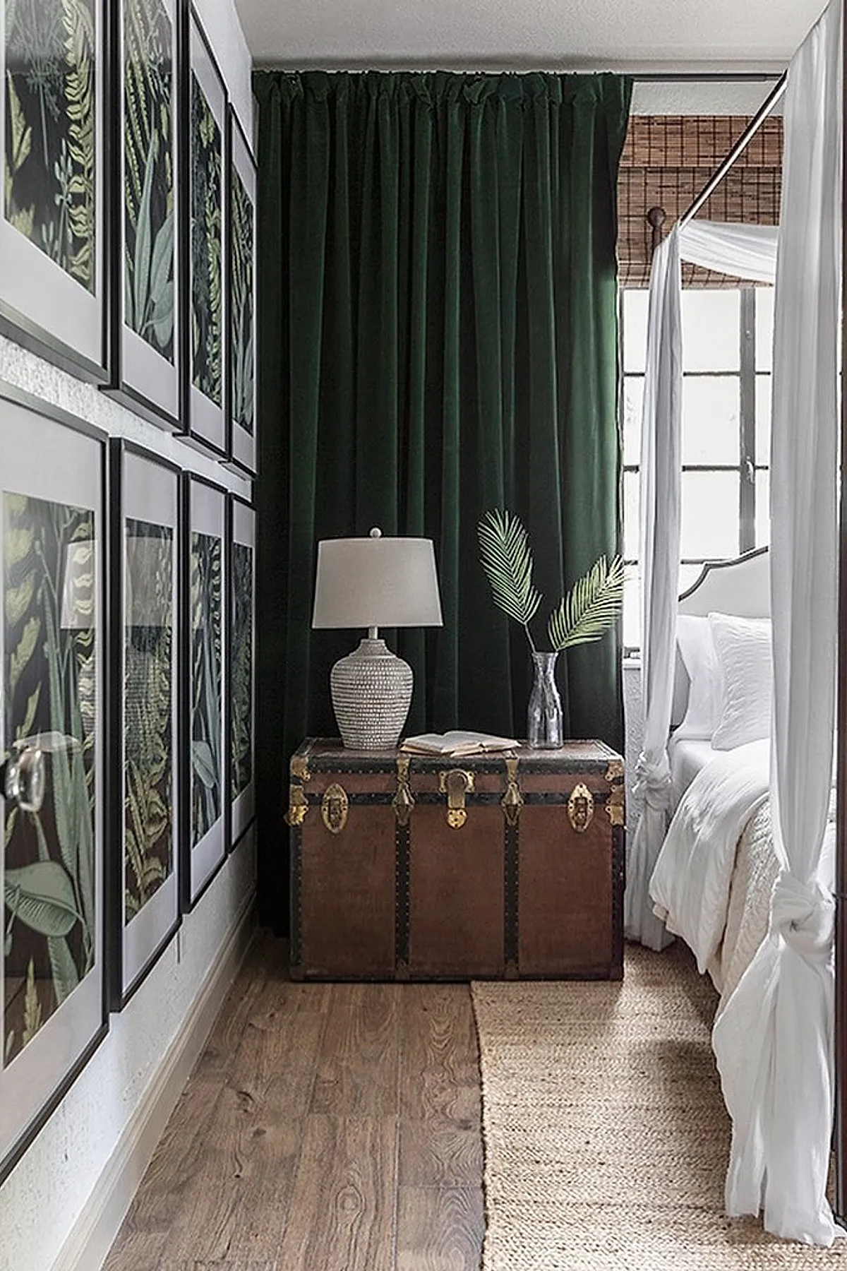 Bedroom corner featuring a vintage brown trunk used as nightstand, dark green velvet curtains, white bed with canopy frame and white drapes, multiple framed botanical prints on wall, ceramic table lamp and glass vase with palm leaves on trunk.