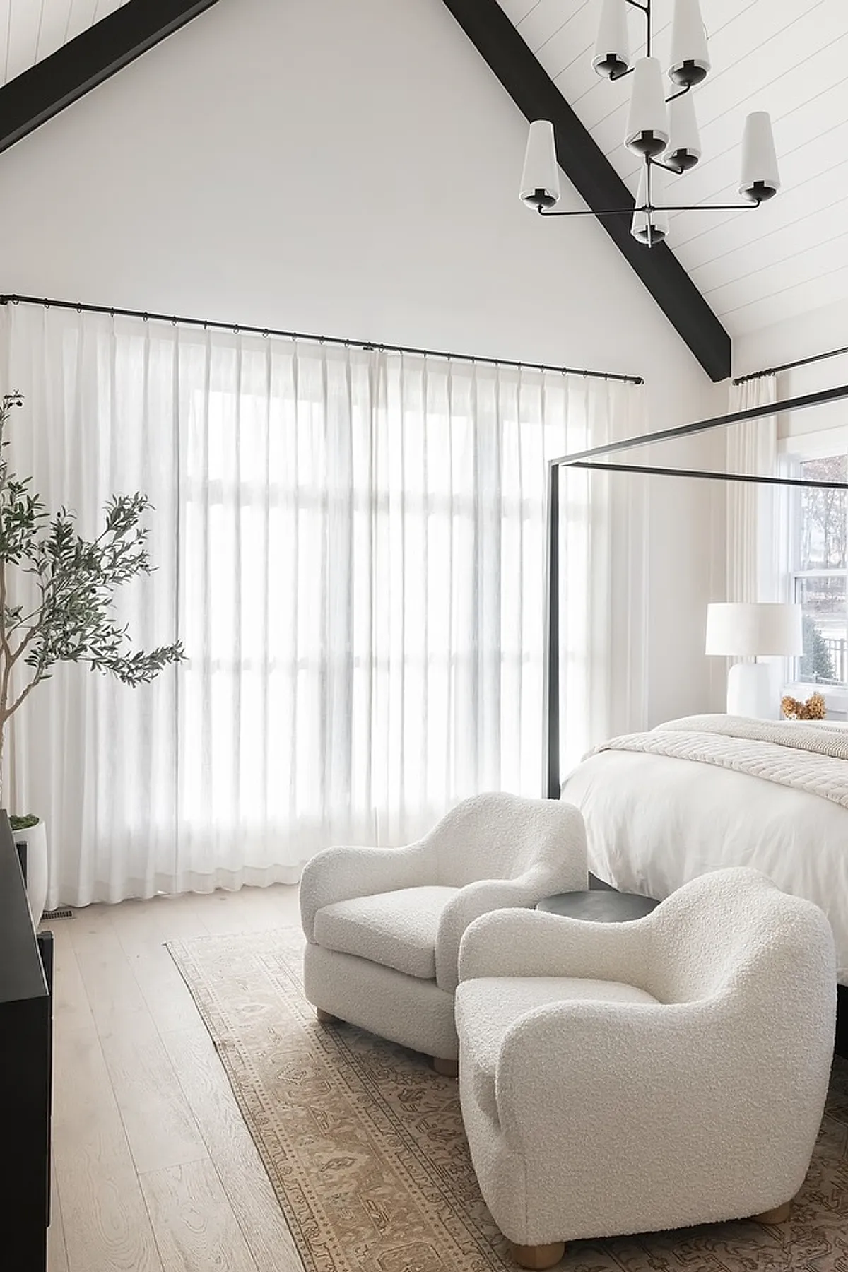 Bright bedroom with vaulted ceiling featuring black metal canopy bed, two curved white textured armchairs on a beige patterned rug, sheer white curtains covering large windows, and light wood floor.