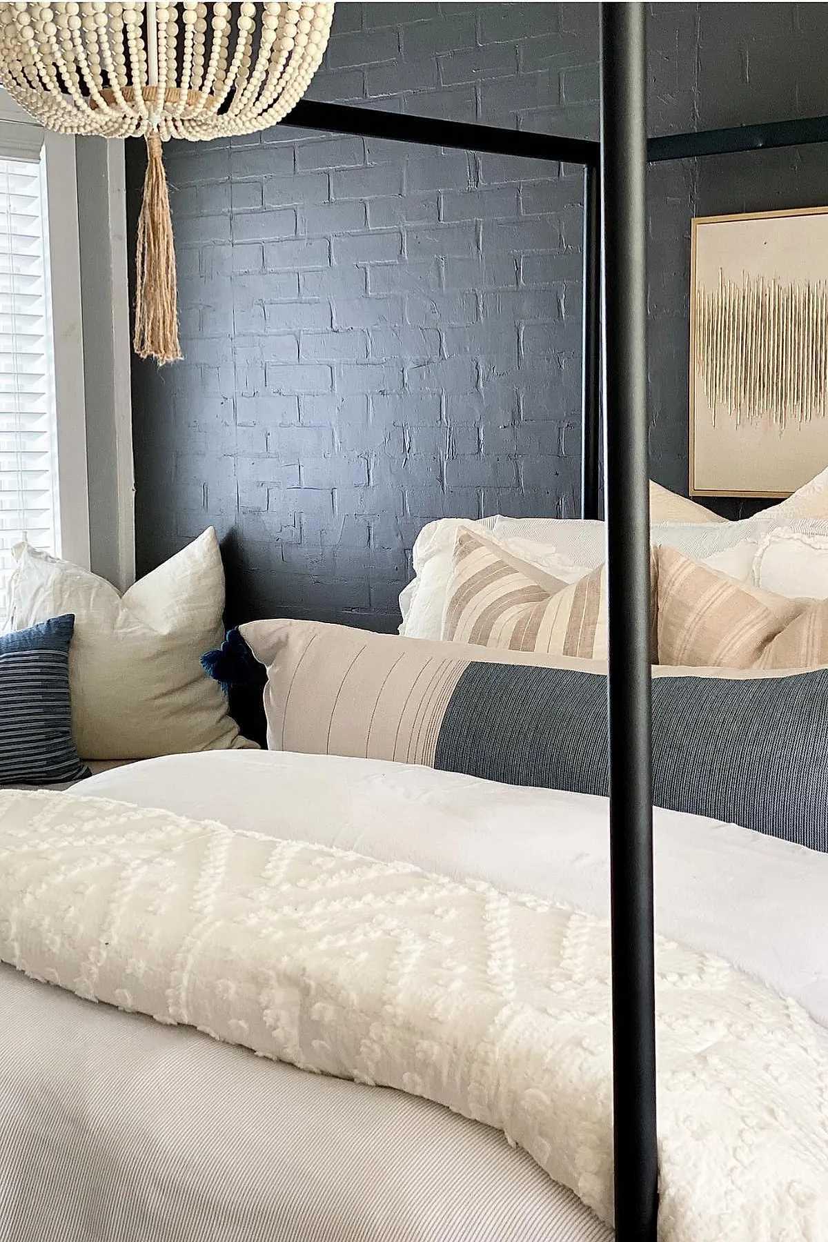 Black metal canopy bed frame against a dark gray textured brick wall, layered with cream, beige, and navy striped pillows, white bedding, and a large natural wood bead chandelier hanging overhead.