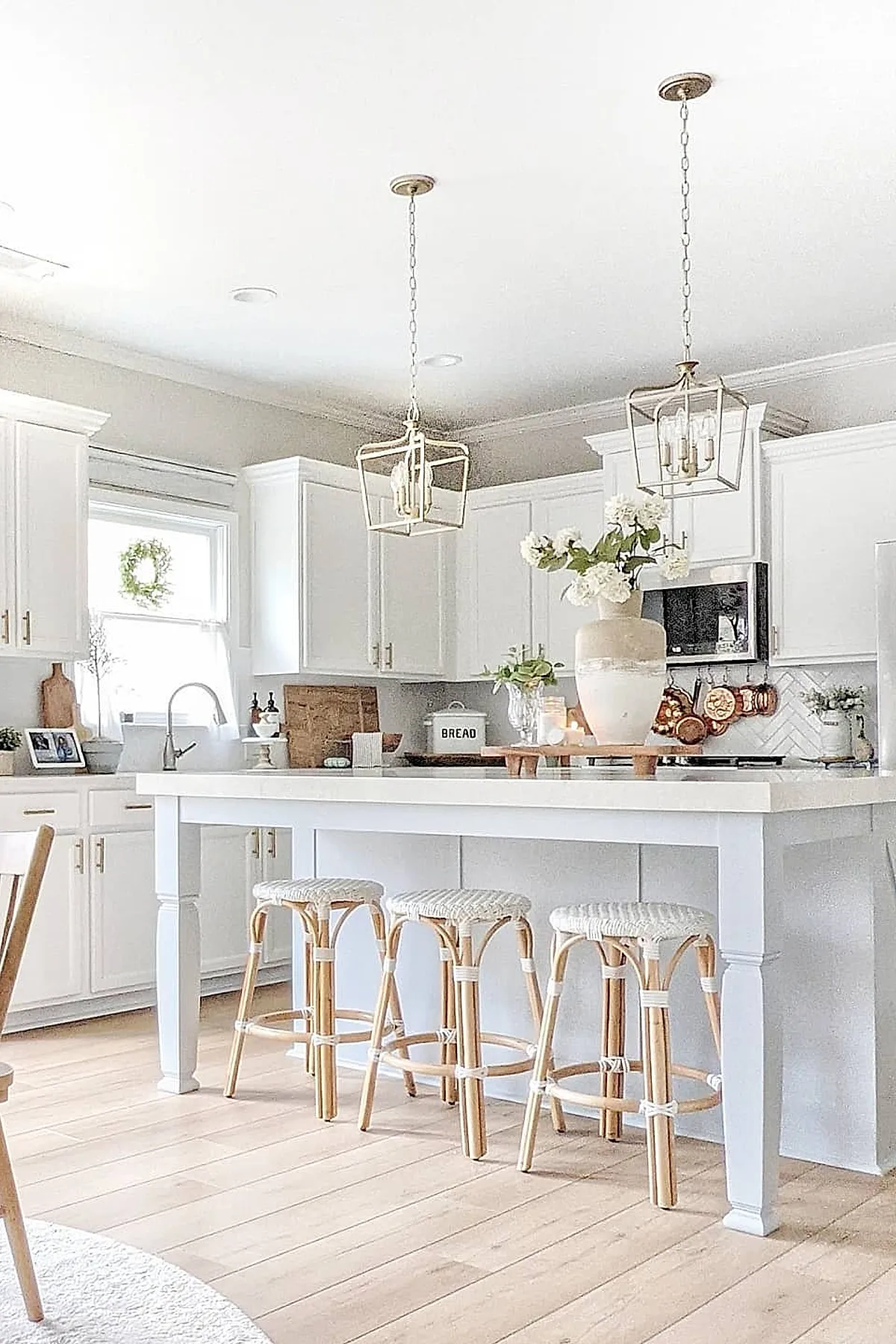 White French country kitchen with large island, natural rattan bar stools with woven seats, brass cage pendant lights, white cabinetry, light wood floor.