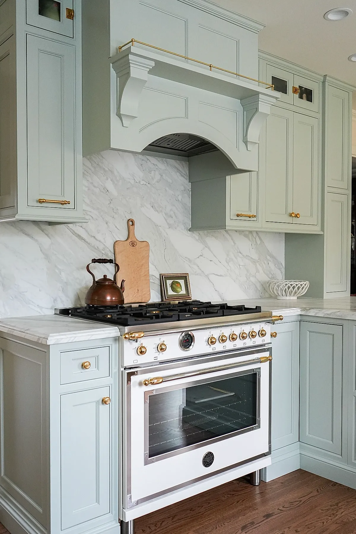 French country kitchen featuring pale green paneled cabinets with gold hardware, white marble backsplash with gray veins, white stove with gold knobs, copper kettle on stovetop, and hardwood floor.