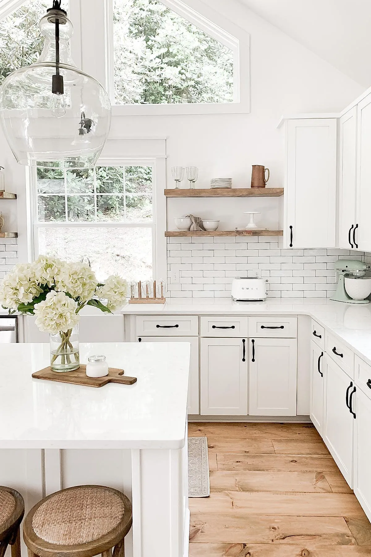 Bright white French country kitchen with black metal cabinet handles, open wooden shelves holding dishes and glassware, white subway tile backsplash, and natural wood floor.