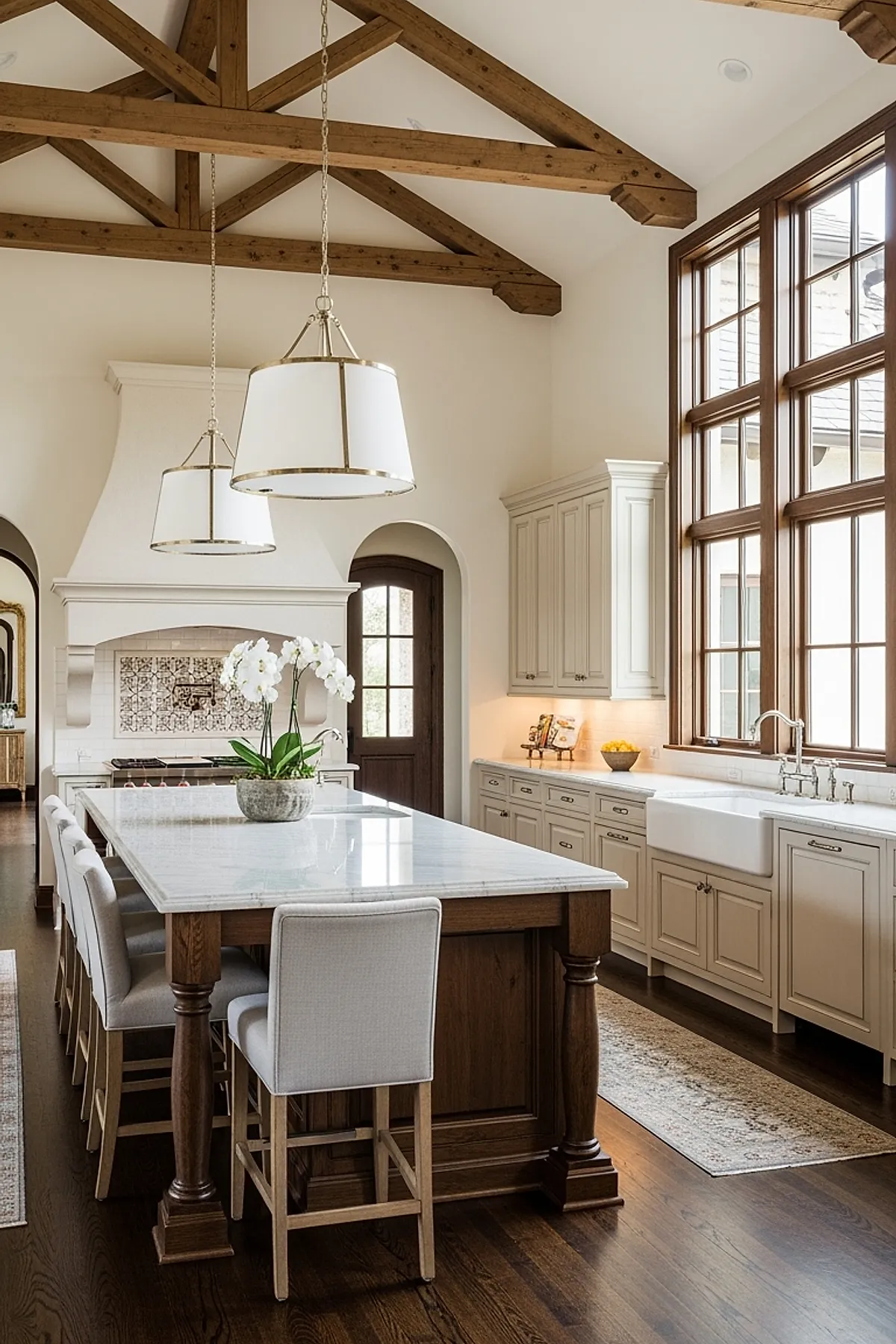 French country kitchen with exposed natural wood ceiling beams, large marble island with wooden base and upholstered stools, white cabinetry with brass hardware, farmhouse apron sink under large windows, and two pendant lights with brass trim.