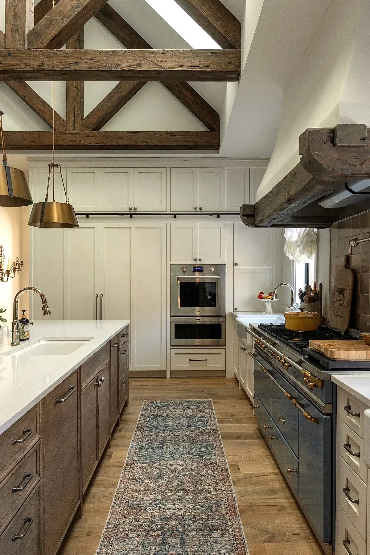 French country kitchen with exposed dark wooden ceiling beams, brass pendant lights, white upper cabinets, wooden lower cabinets island, patterned runner rug on wood floor.