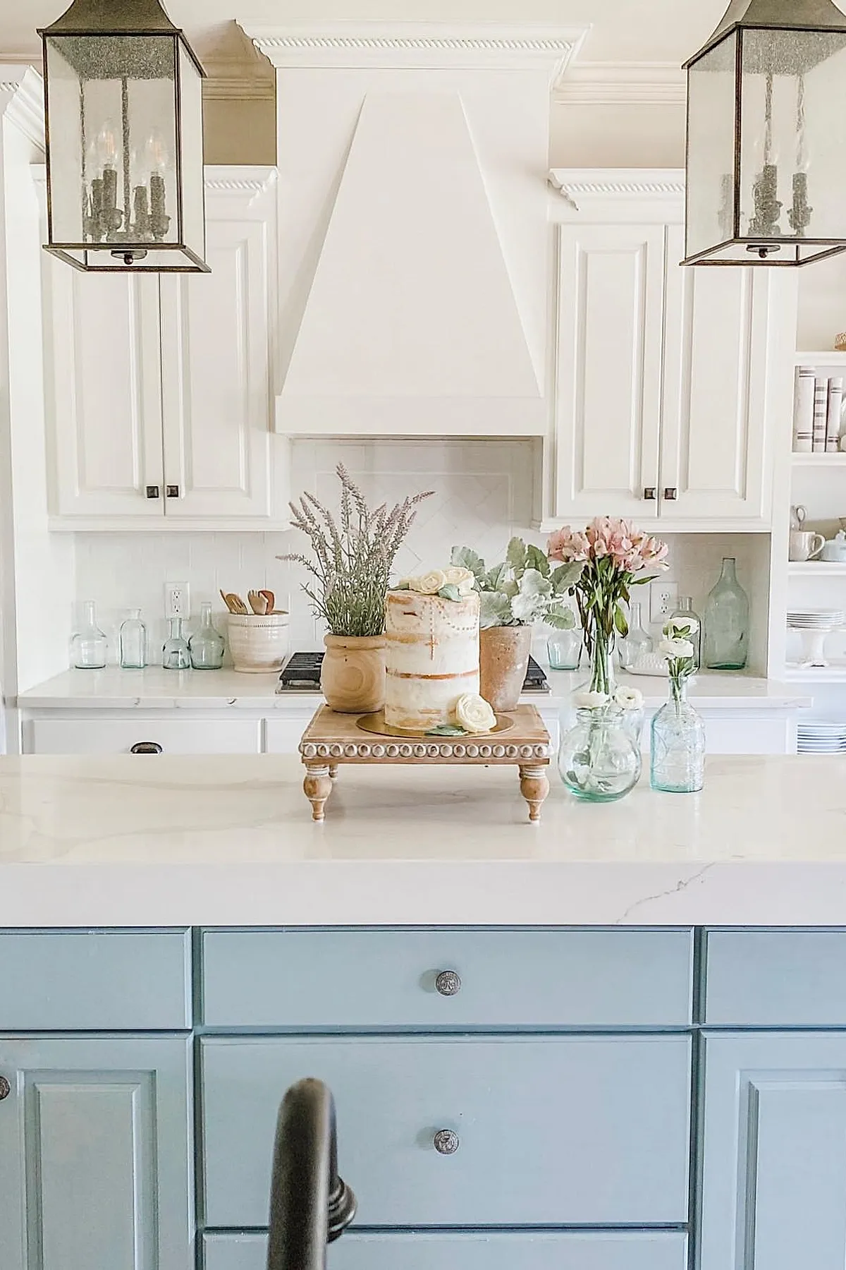 French country kitchen with pale blue island cabinets, white upper cabinets and range hood, marble countertop adorned with wooden pedestal holding potted plants and flowers, clear glass vases with fresh flowers.
