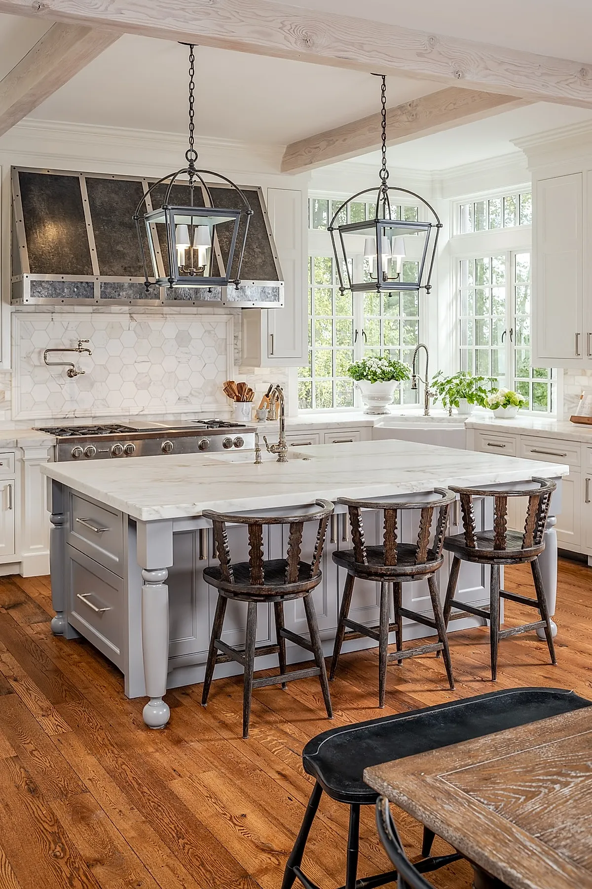 French country kitchen featuring a large gray painted island with white marble countertop, dark rustic wooden chairs, metal industrial-style range hood, white cabinetry, hexagonal tile backsplash, exposed wooden ceiling beams, hardwood floors.