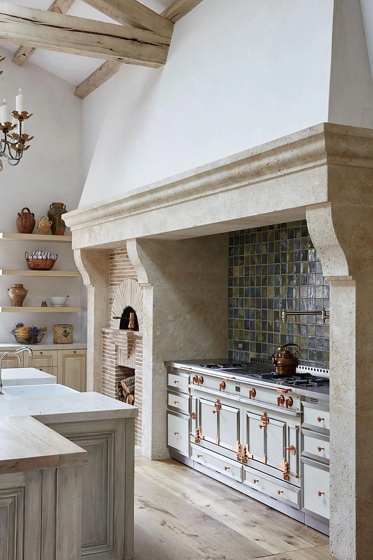 French country kitchen with large stone arch over stove, gray and green square tiled backsplash, white cabinetry with copper hardware, light wood floors, exposed wooden ceiling beams.
