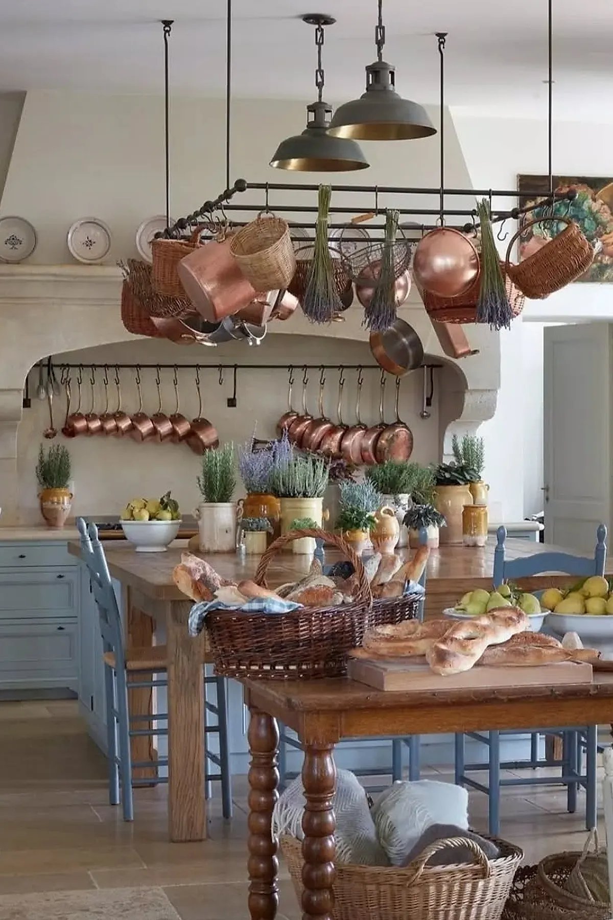 French country kitchen with copper pots and pans hanging above wooden tables, dried lavender bundles, wicker baskets, blue painted chairs, ceramic herb pots, and bread baskets on tables.