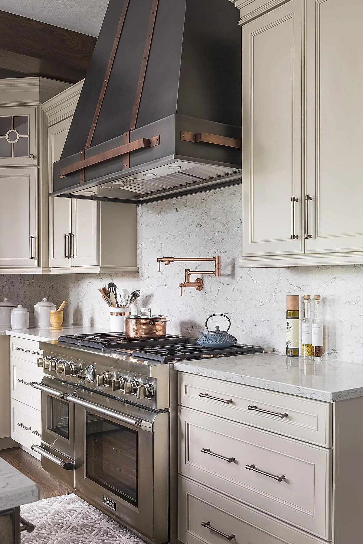 French country kitchen featuring large black metal range hood with copper straps, stainless steel gas stove, copper pot filler faucet mounted on light marble backsplash, cream cabinetry with dark handles, wooden cooking utensils in white ceramic holder.