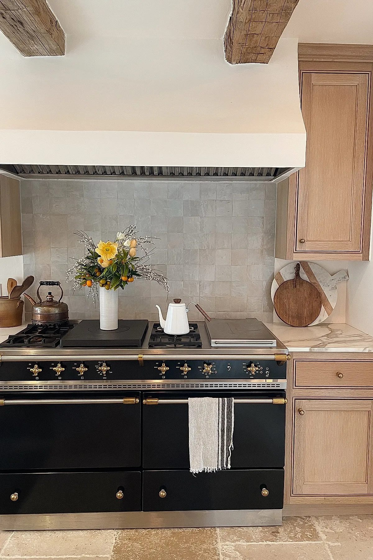French country kitchen featuring a black gas range with brass hardware, light wood cabinets, marble countertop, beige tiled backsplash, wooden cutting boards on counter, and fresh yellow and orange flowers in white vase on stove.