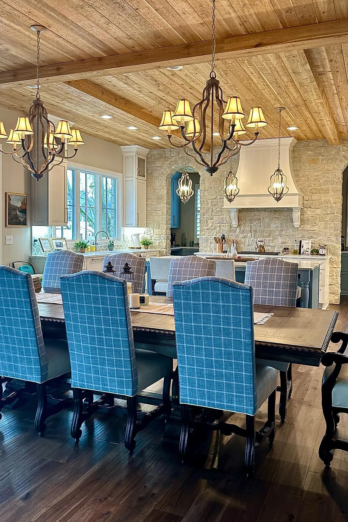 French country kitchen with blue and gray plaid upholstered dining chairs, wooden dining table, exposed natural wood plank ceiling with beams, wrought iron chandeliers with lampshades, stone wall and white cabinetry in background.