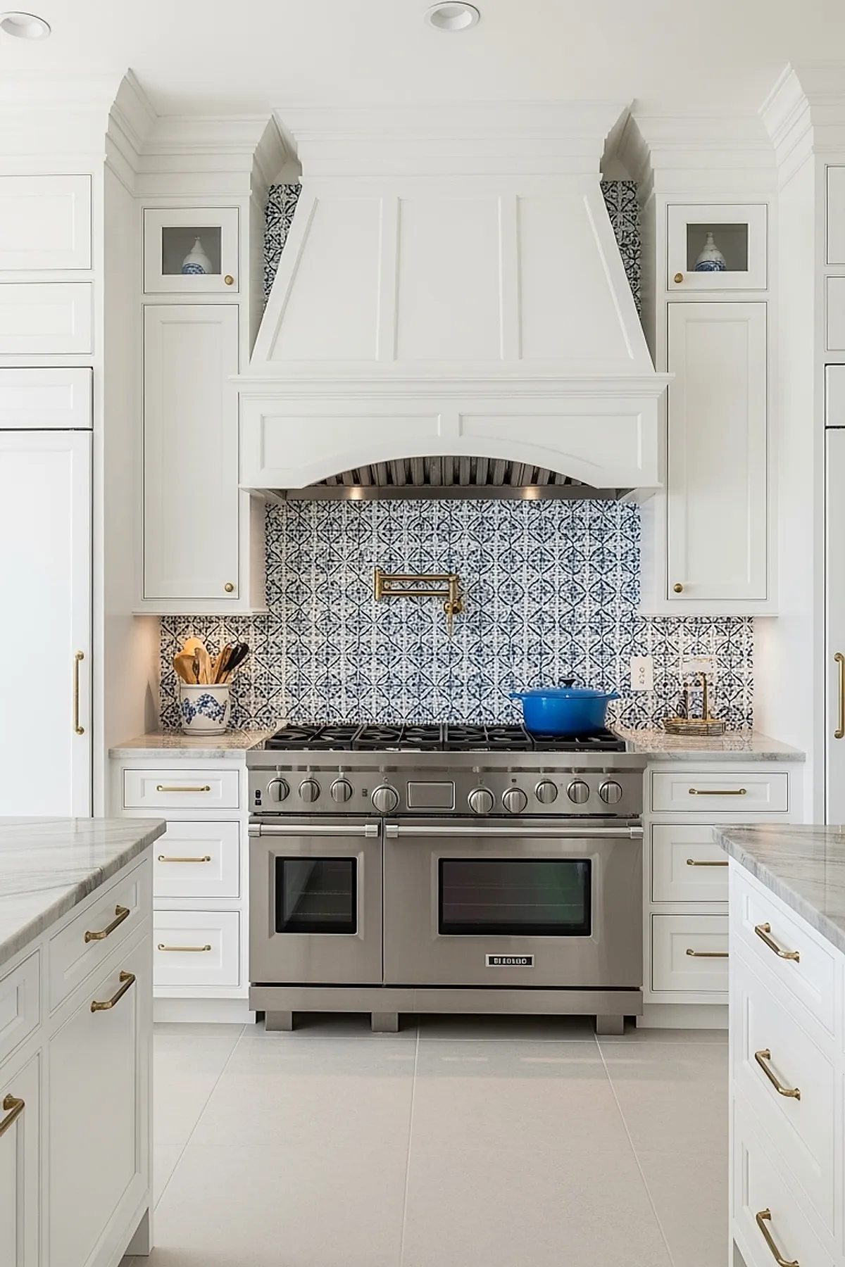 French country kitchen featuring white cabinetry, blue and white patterned tile backsplash, stainless steel range with double oven, brass pot filler above stove, and brass cabinet hardware.