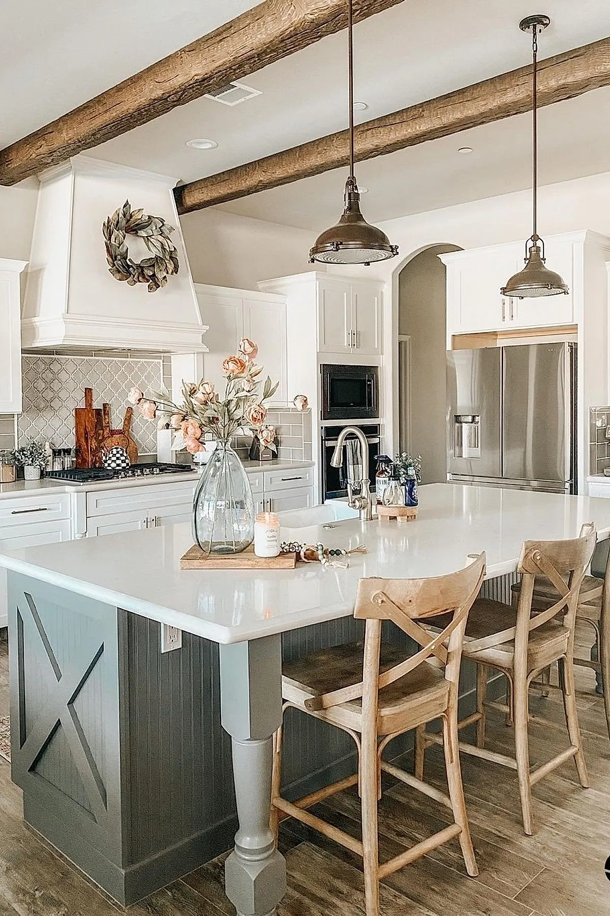 French country kitchen featuring large island with gray painted base and white countertop, exposed natural wood ceiling beams, bronze pendant lights, wooden cross-back stools, white cabinetry, stainless steel appliances.