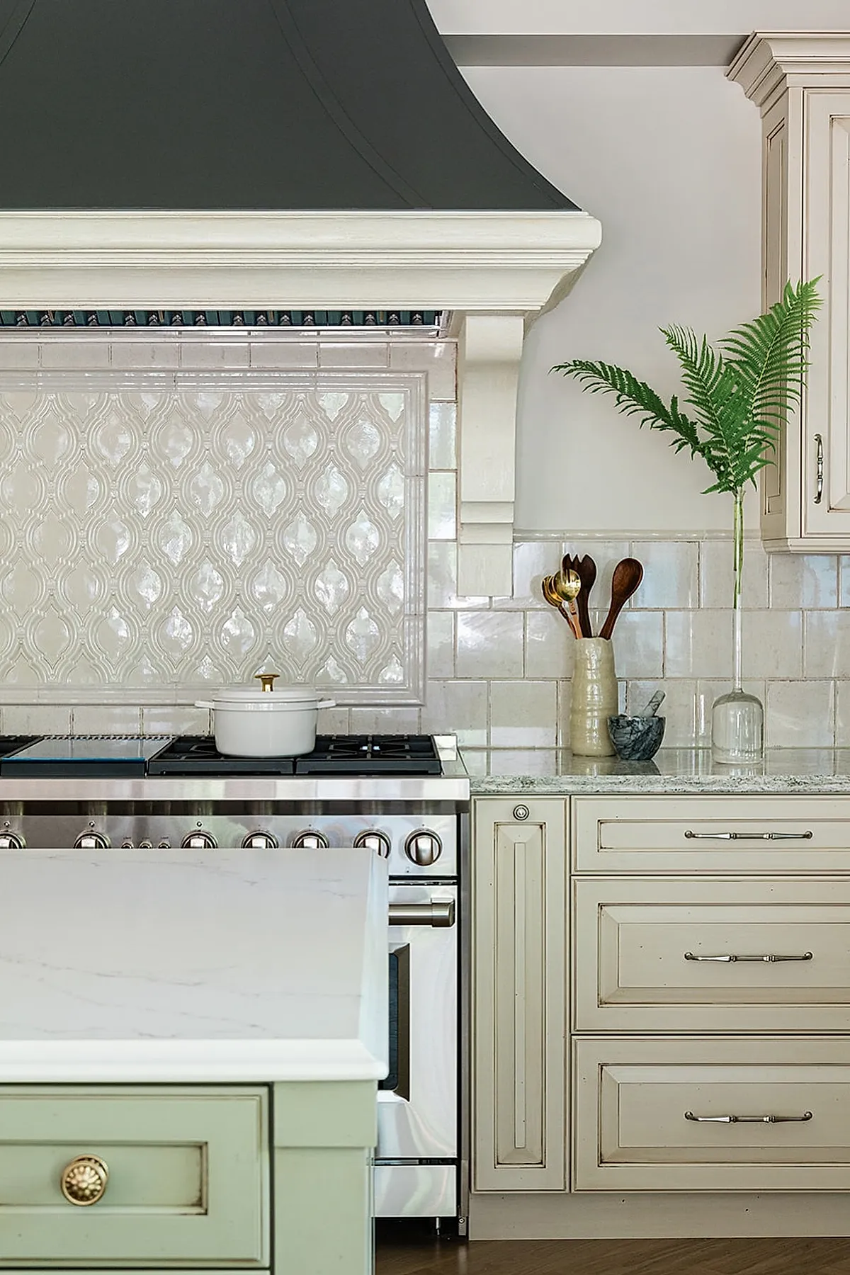 French country kitchen with cream-colored textured tile backsplash, stainless steel gas stove, pale cabinetry with silver handles, granite countertop, green island with brass knob, and glass vase holding fern leaves.