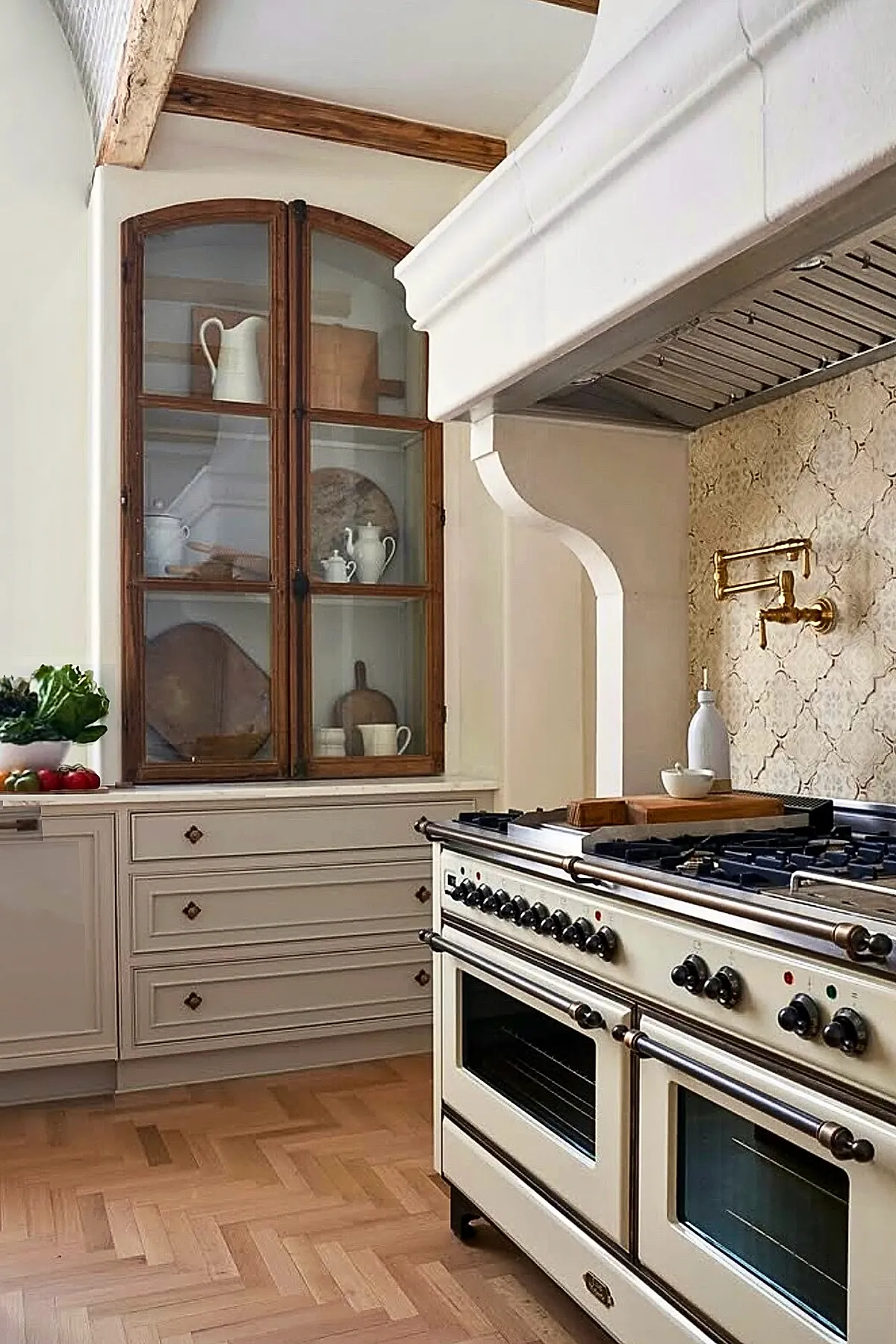 French country kitchen featuring cream-colored stove with brass pot filler, beige patterned tile backsplash, arched wooden glass cabinet displaying white pitchers and pottery, exposed wooden ceiling beams, light hardwood floor.