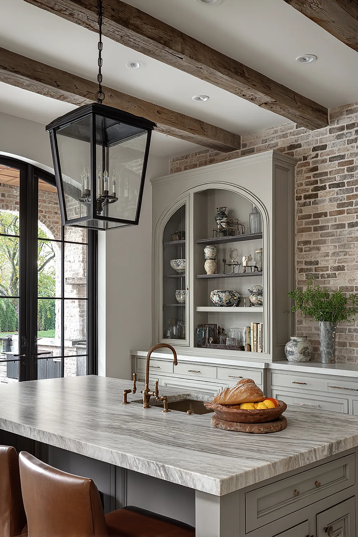 French country kitchen with large marble island featuring brass faucet, exposed rough wood ceiling beams, light gray cabinetry, glass-front cabinet with ceramics, brick walls, and leather chairs.