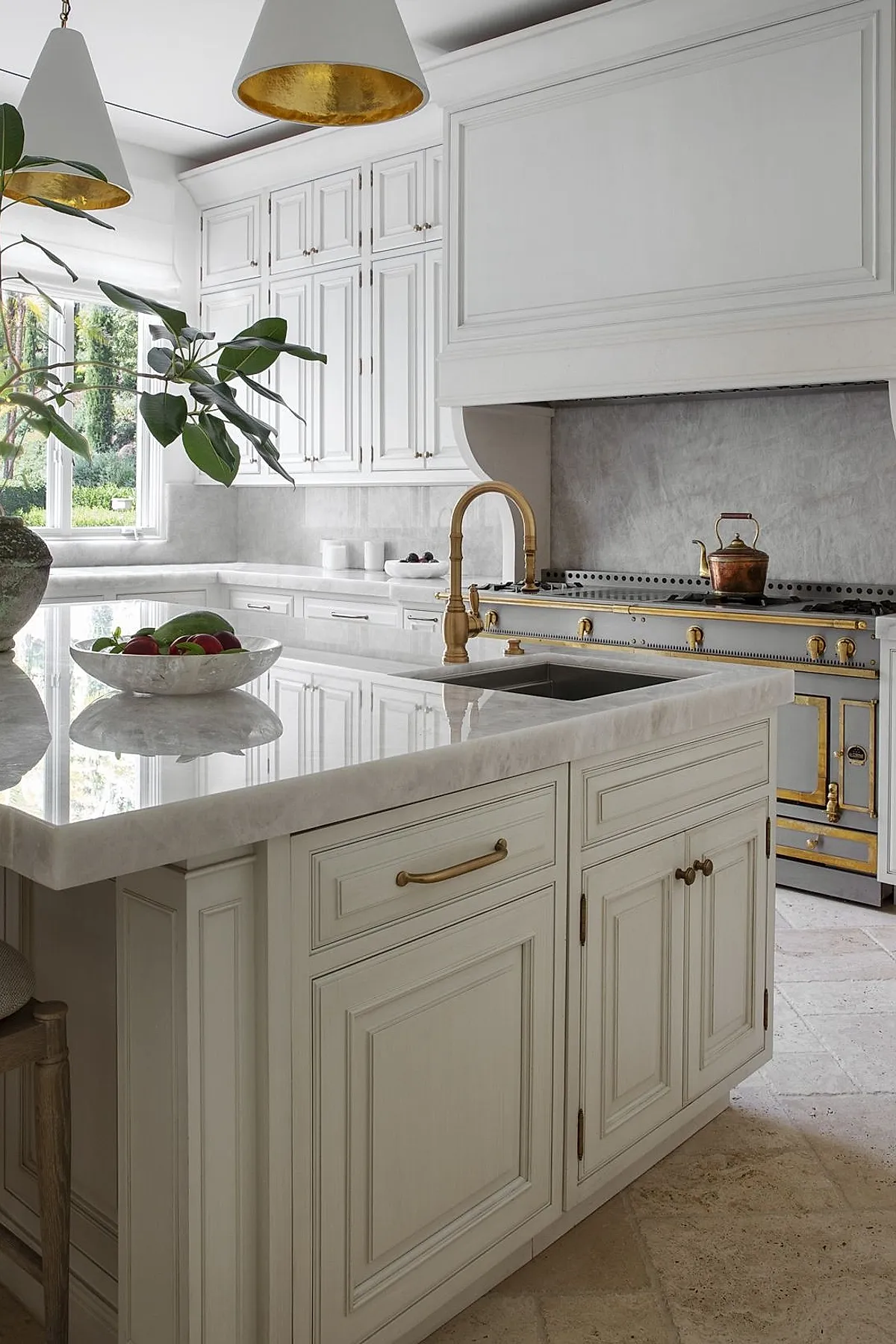 French country kitchen featuring white paneled cabinets, brass faucet, polished marble countertops, light gray stone backsplash, vintage stove with brass accents, and large windows overlooking greenery.
