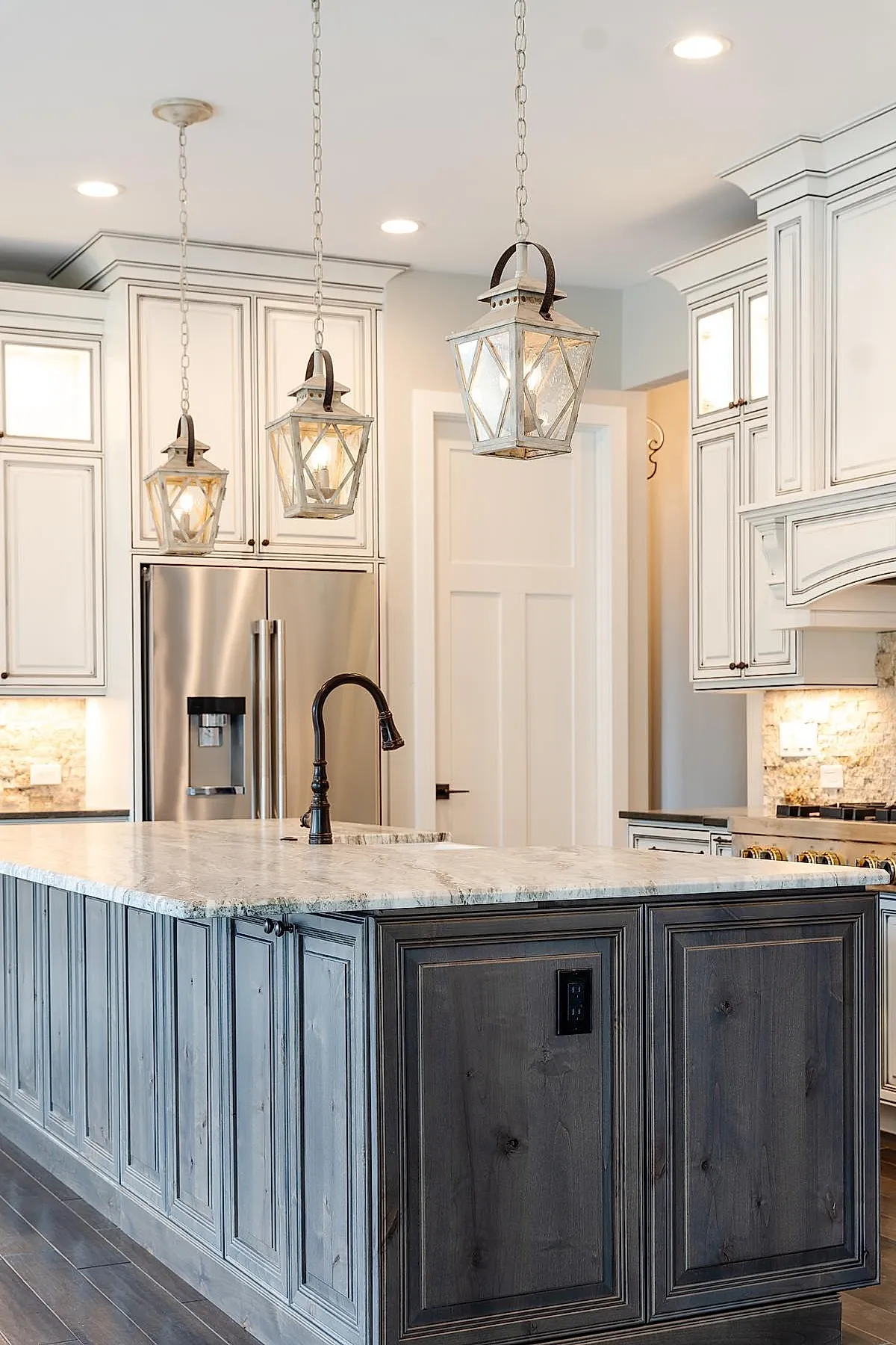 French country kitchen featuring a large gray wooden island with marble countertop, three glass lantern pendant lights hanging above, bronze faucet, white cabinetry, and stainless steel refrigerator.