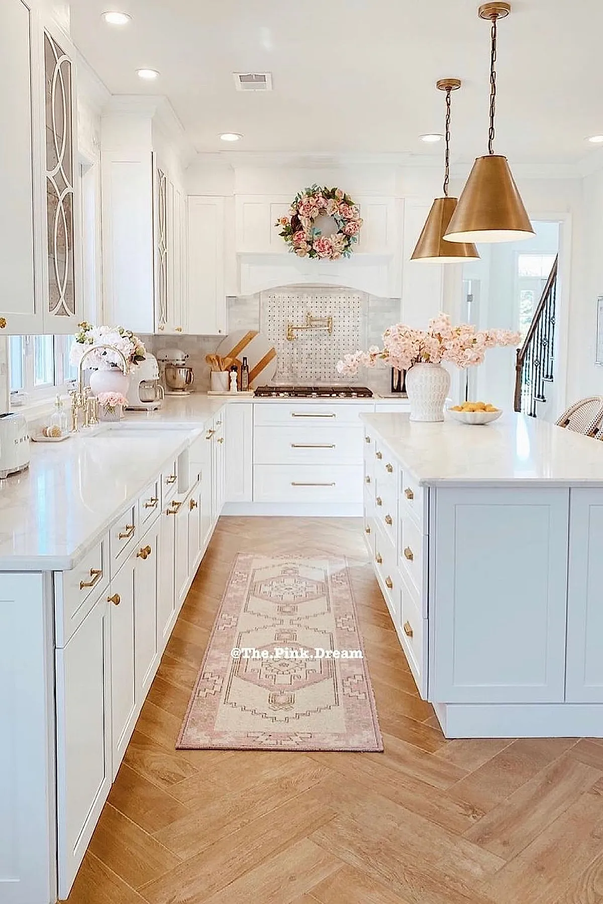 Bright white French country kitchen featuring gold cabinet hardware, white marble countertops, brass faucet, pink floral wreath above stove, gold pendant lights, light wood herringbone floor, and pink flower arrangements on counters.