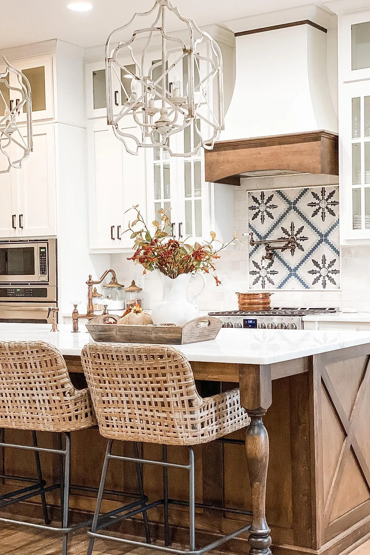 French country kitchen featuring woven rattan bar stools, wooden kitchen island with turned legs, white cabinetry, decorative blue and gray patterned tile backsplash, white range hood with wood trim, and floral centerpiece in white pitcher on island.