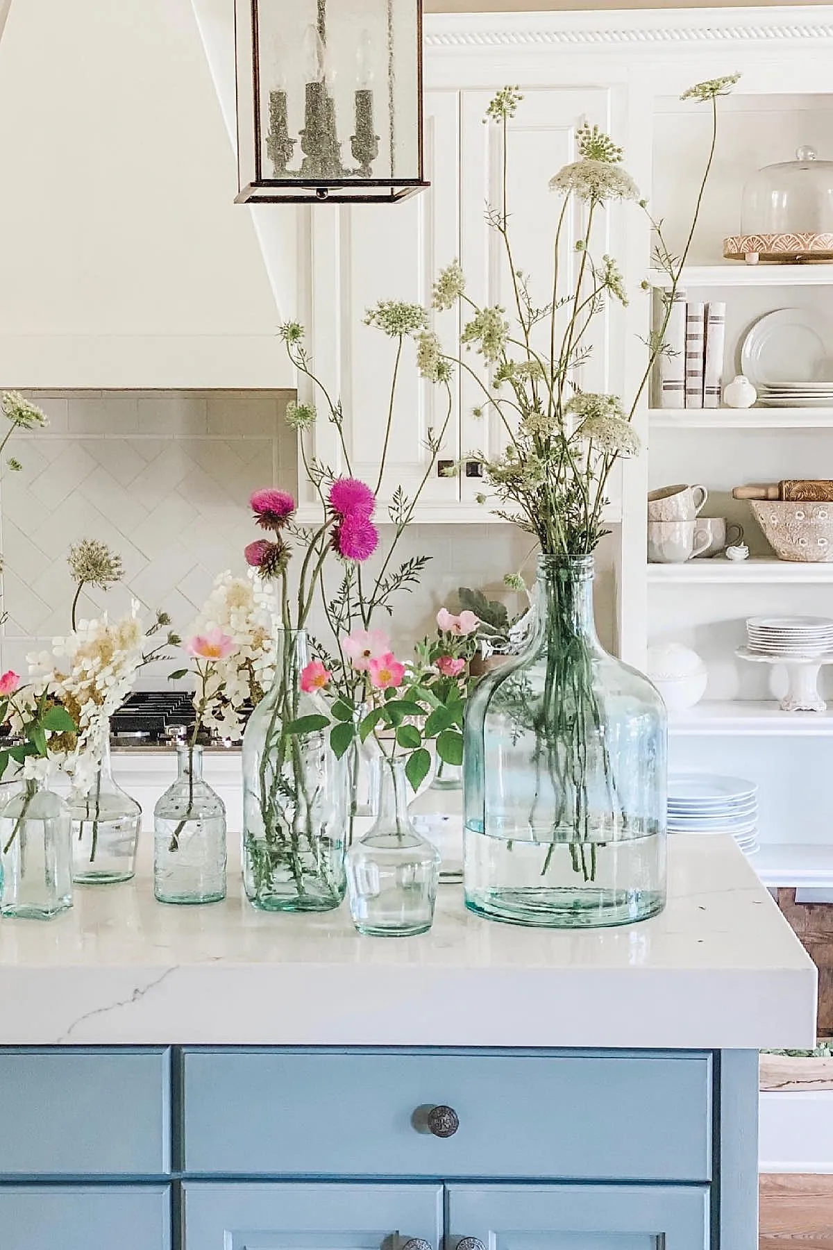 French country kitchen featuring blue painted island with white marble countertop, various clear glass bottles holding wildflowers including pink and white blooms, white cabinetry and open shelving in background.