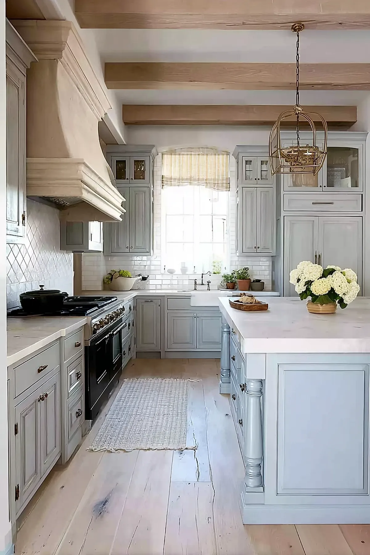 French country kitchen with soft blue painted cabinets, large white marble island, exposed natural wood ceiling beams, white tiled backsplash, black stove, and light hardwood floors.