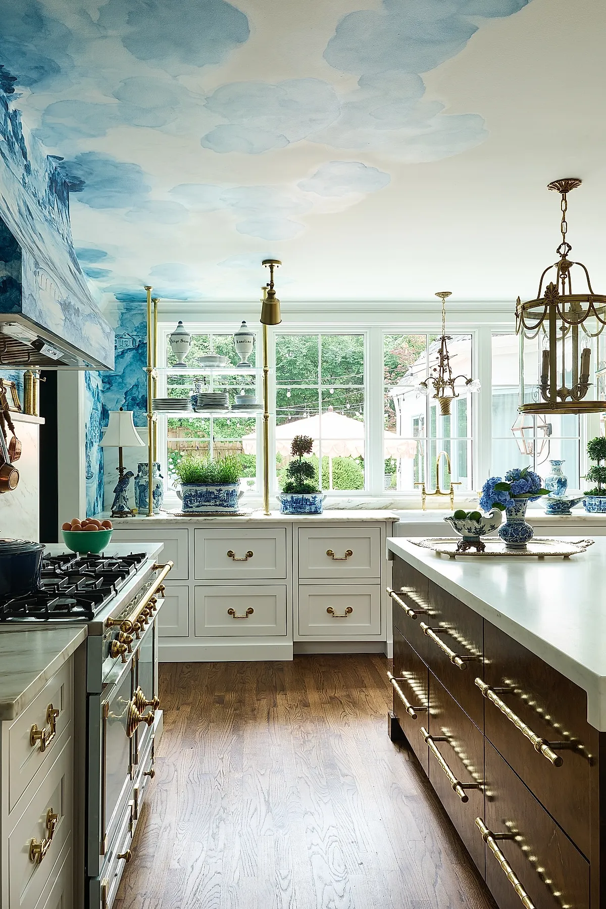 French country kitchen with white cabinets, brass hardware, blue hand-painted cloud ceiling mural, blue-and-white pottery, large windows, wooden floor.