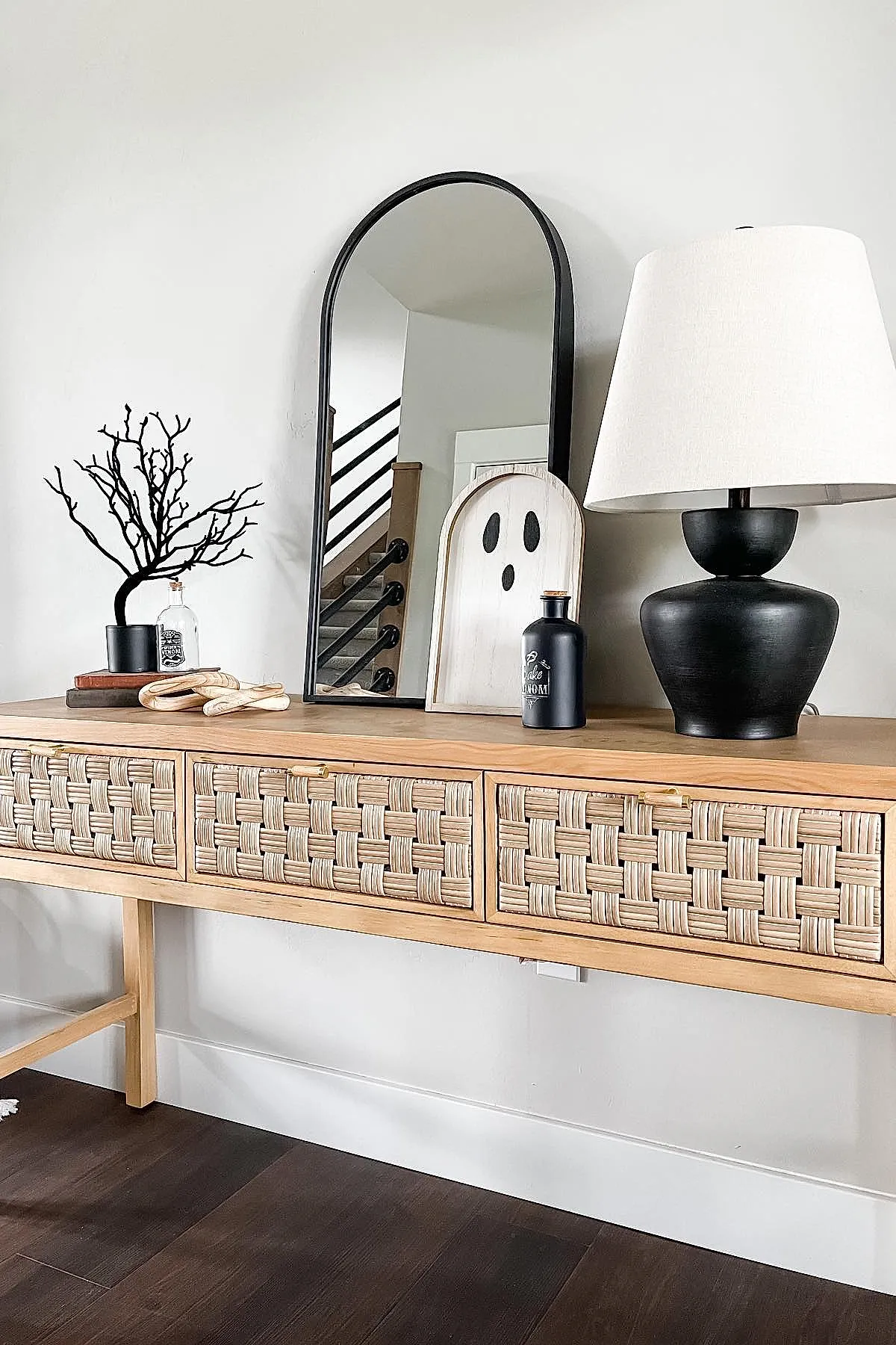 Light wood console table with woven drawer fronts holding a black potted branch sculpture, stacked books, white wooden ghost sign leaning on black-framed arched mirror, matte black bottle and lamp with white shade against a white wall.