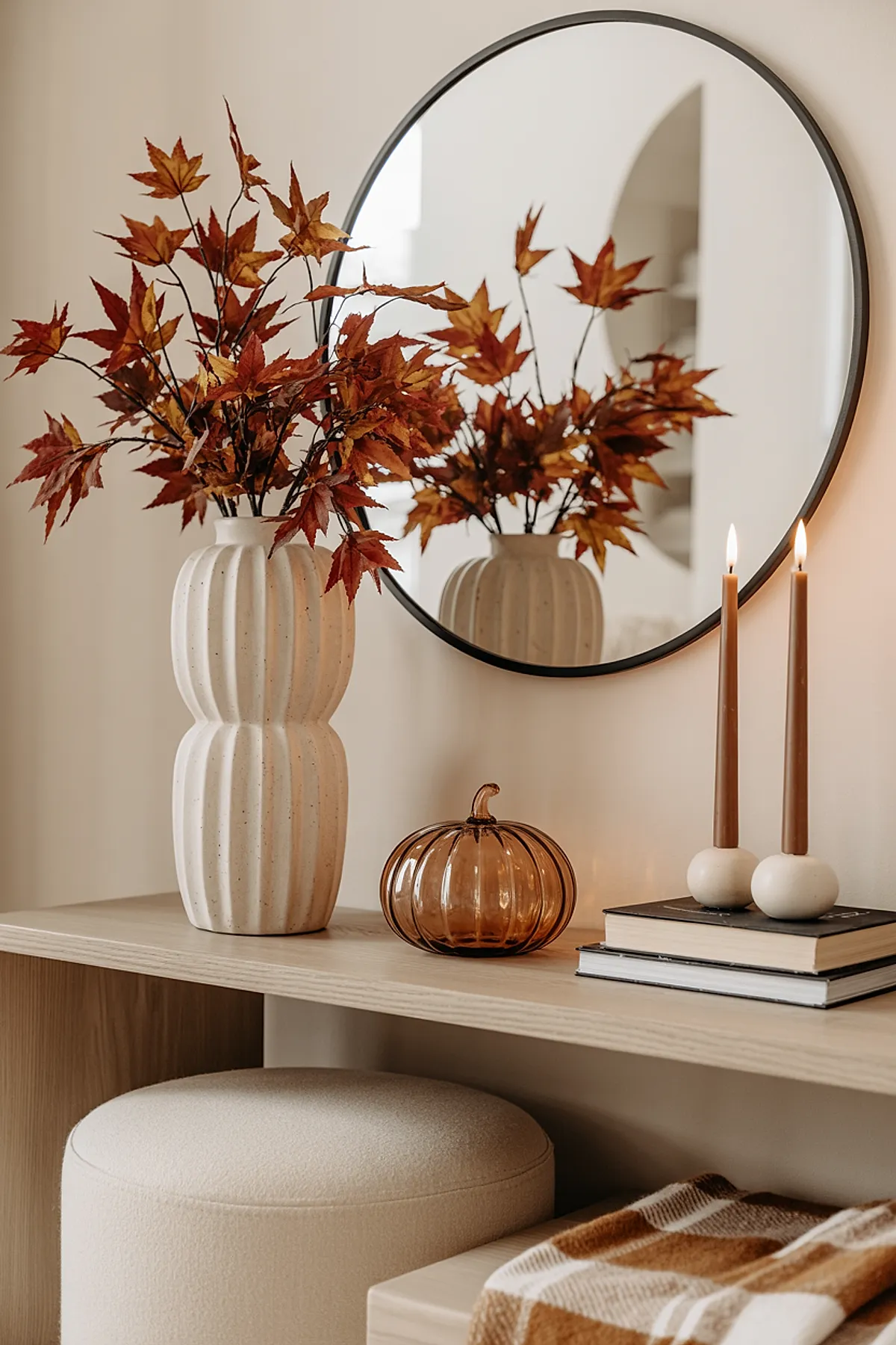 Light wood console table featuring a tall cream ceramic vase filled with orange and rust autumn leaves, an amber ribbed glass pumpkin, two lit brown taper candles in round white holders atop stacked books, all reflected in a large round black-framed mirror against beige wall.