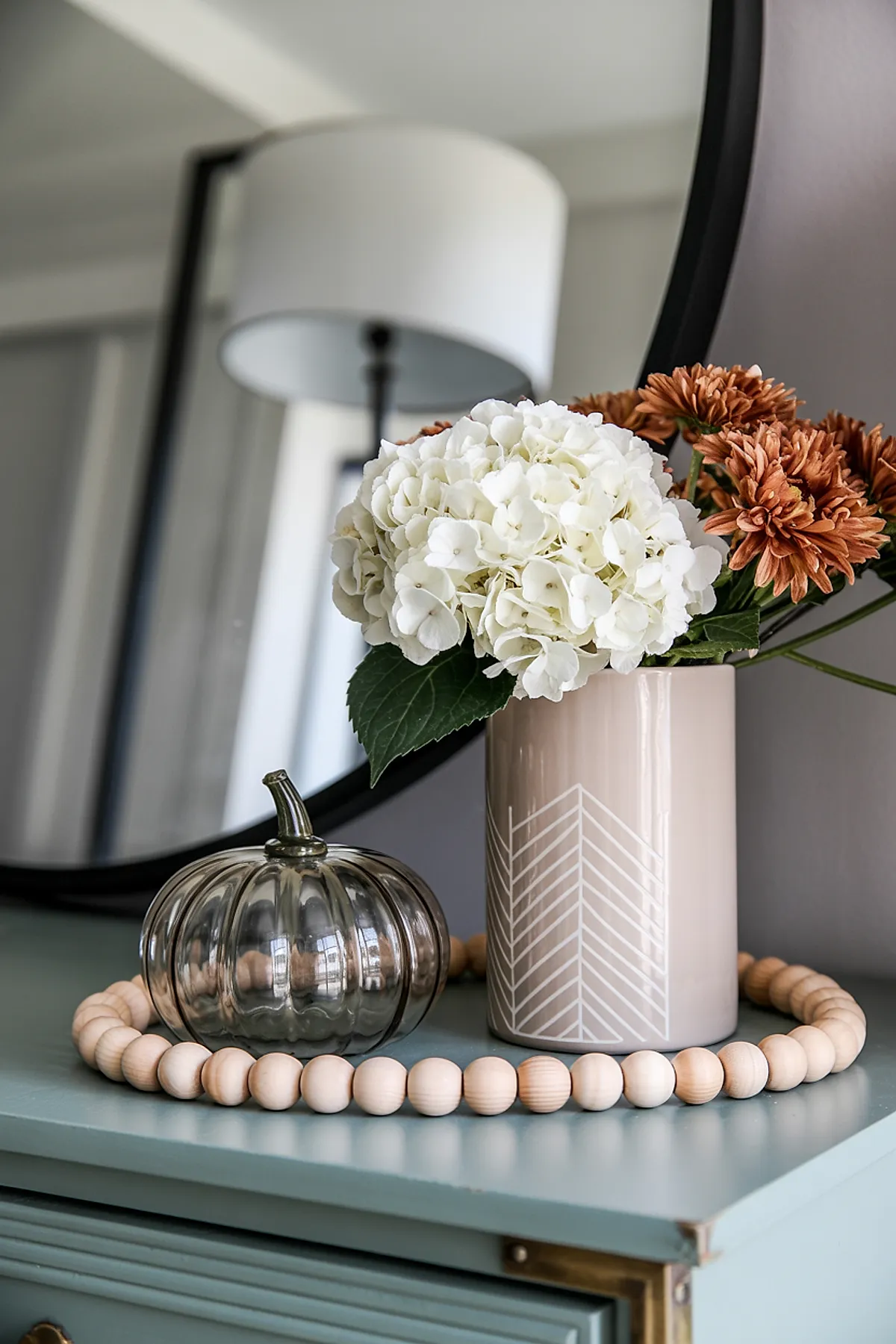 Neutral beige vase holding white hydrangea and muted orange chrysanthemums next to a small smoky glass pumpkin on a blue dresser topped with wooden bead garland, in front of a black-framed mirror.